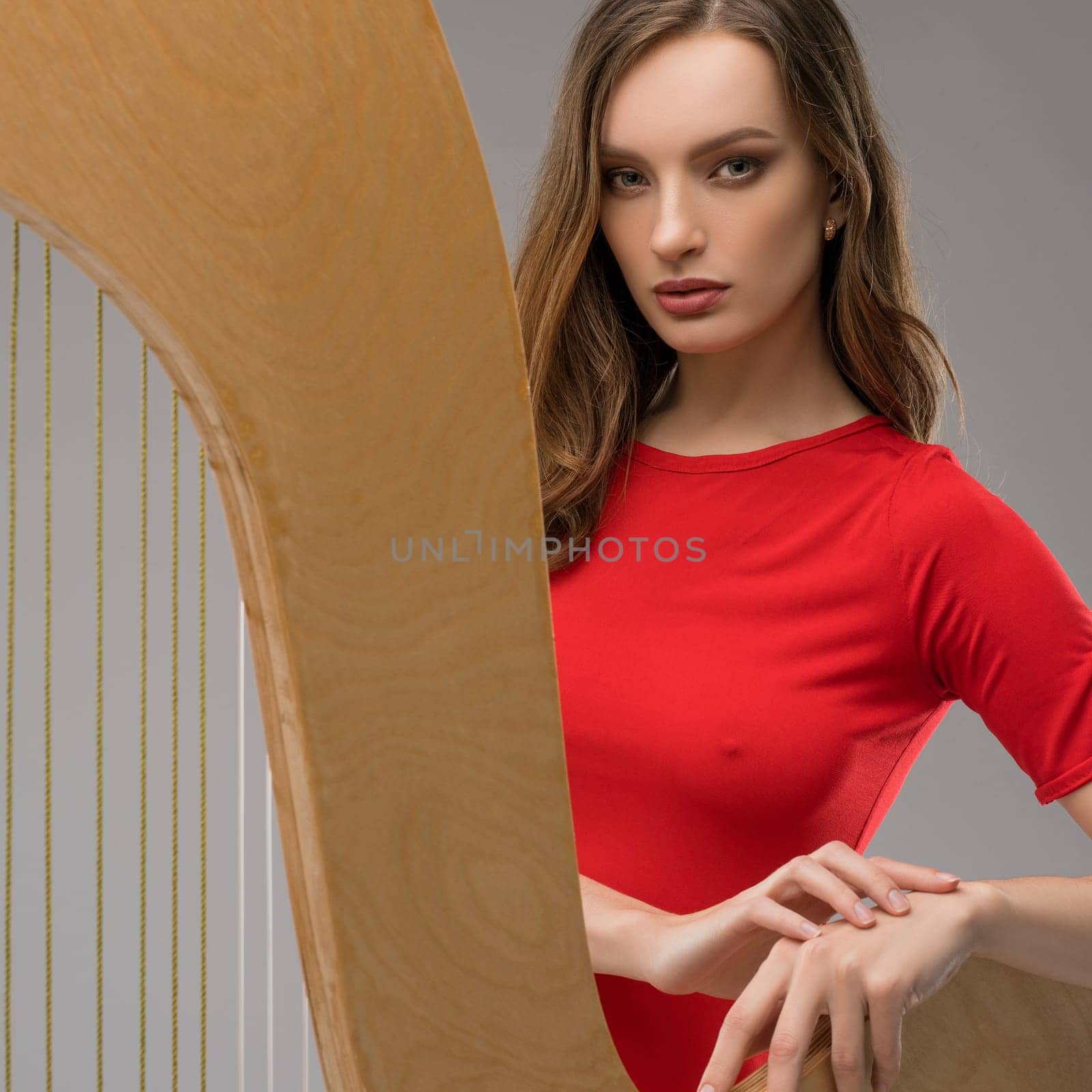 A model dressed in red poses gracefully beside a harp in a professional studio, creating an artistic and elegant atmosphere.