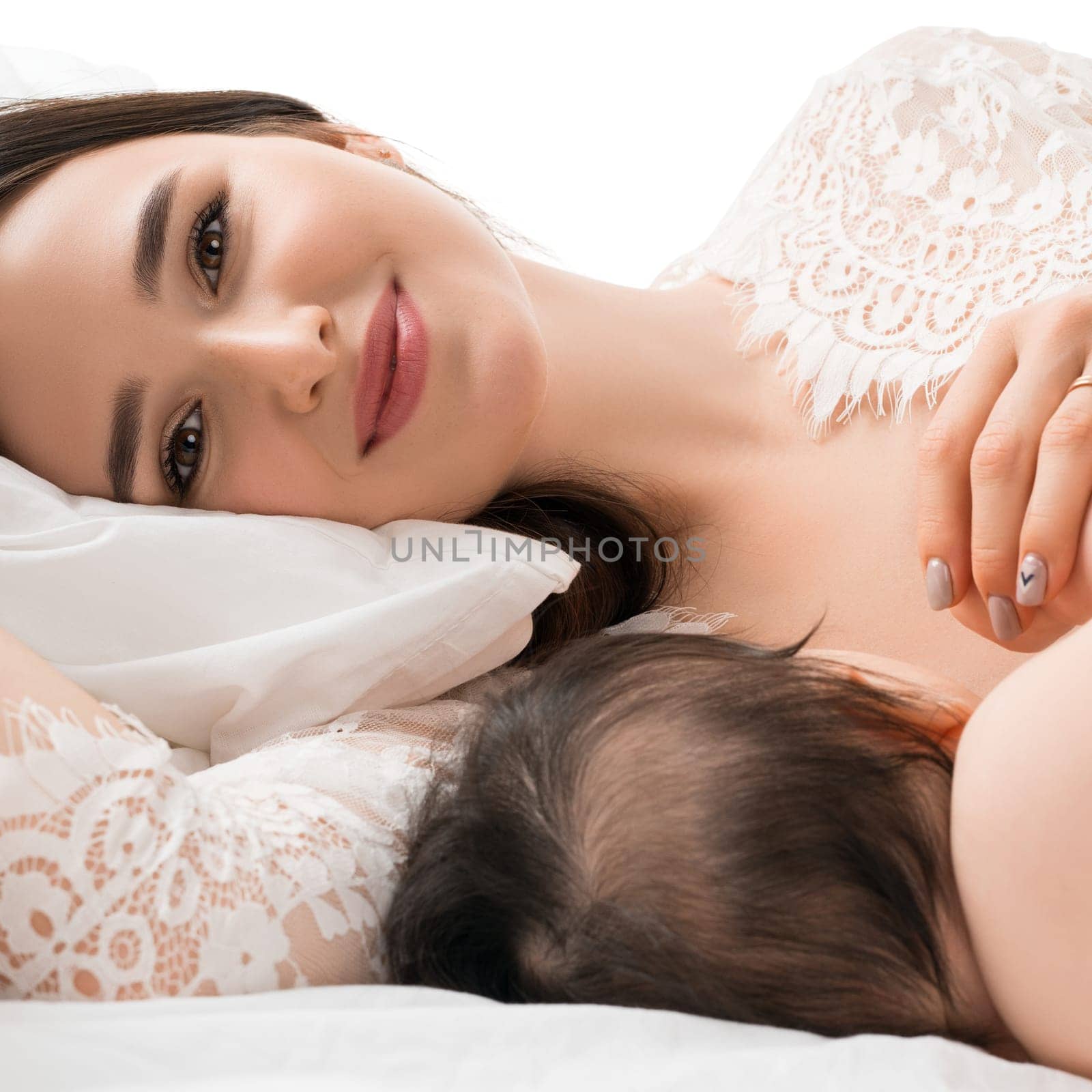 A mother lovingly breastfeeds her infant while lying on a soft bed, surrounded by white pillows and a peaceful atmosphere.