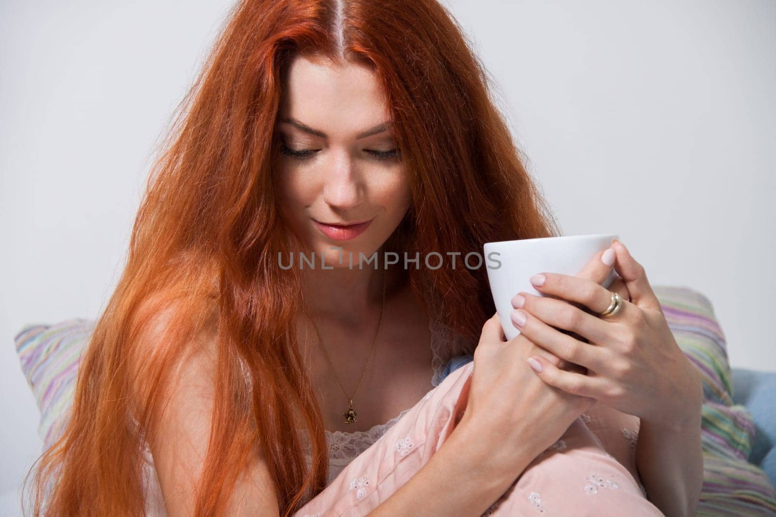 Young red-haired woman sits on her bed with a cup of coffee in the morning after getting up