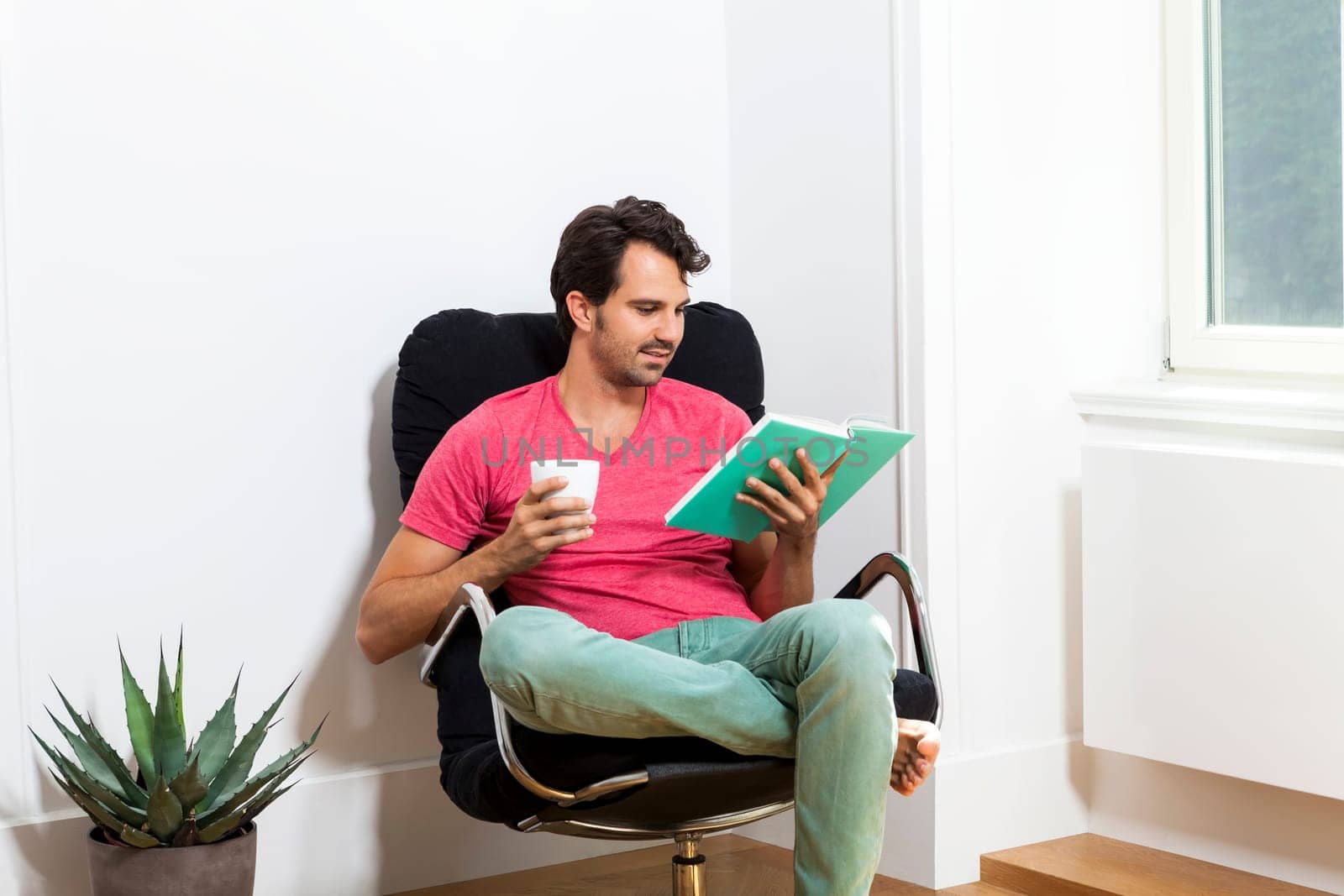 Young man in casual clothes relaxes while reading in an armchair with a drink in his hand