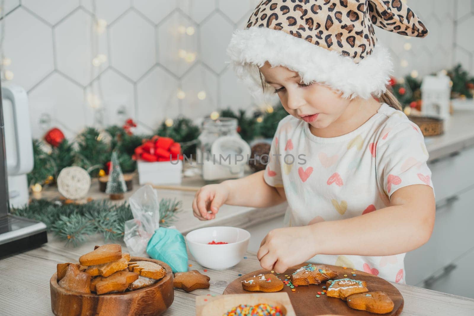 Little dark-haired girl 3 years old in Santa spotted cap decorate gingerbread cookies with icing in white Christmas decorated kitchen. Merry Christmas, Happy New Year