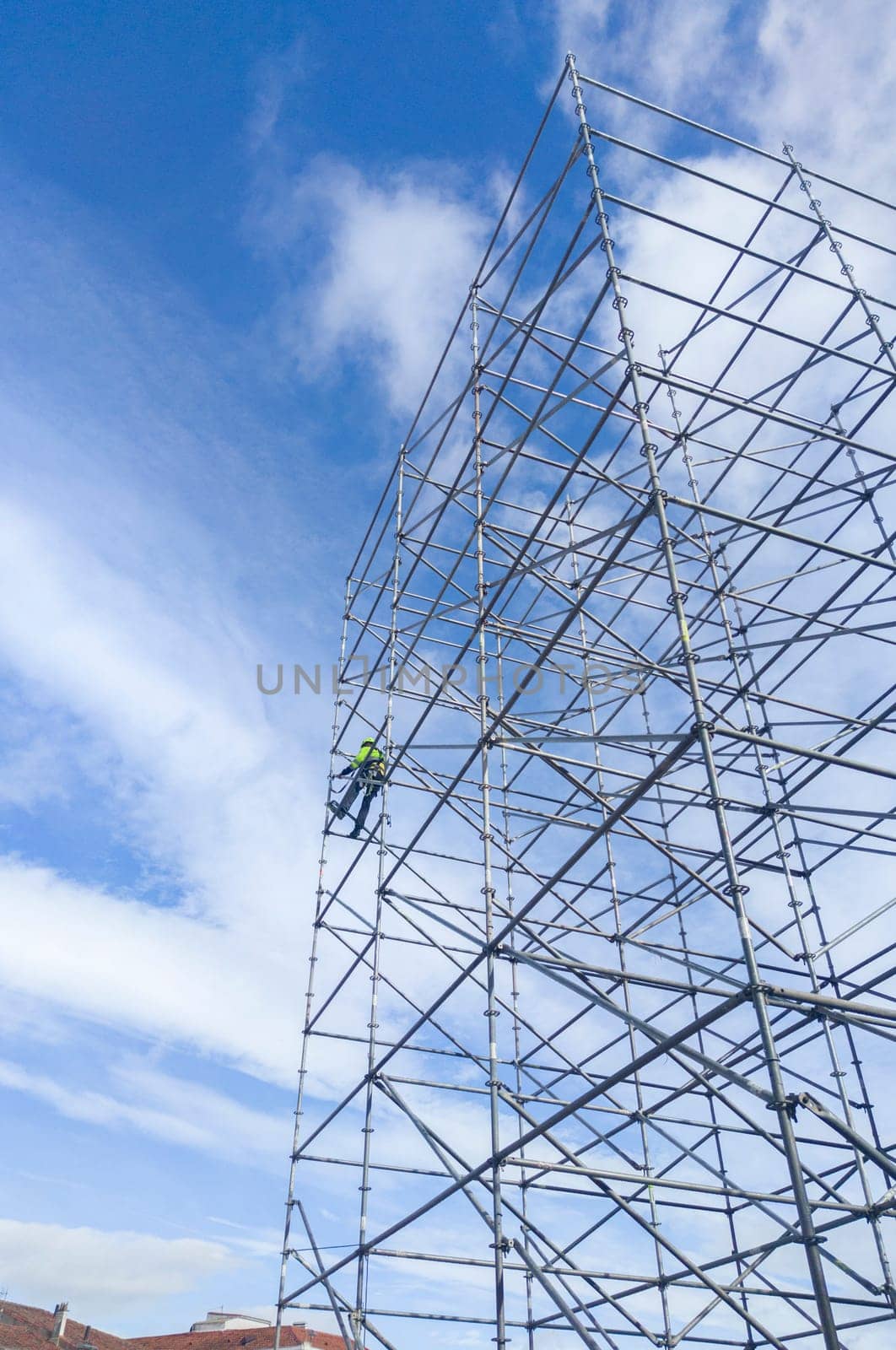 Unidentified worker at height dismantling a scaffolding tower. Blue cloudy sky on background