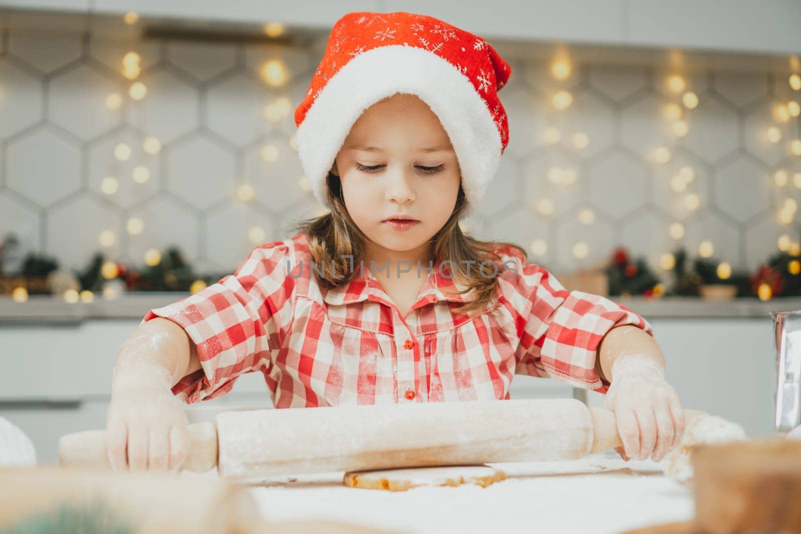 Little dark-haired girl 3 years old in red Christmas cap and checkered shirt roll out dough for gingerbread cookies in white Christmas decorated kitchen with garland lights. Merry Christmas, New Year