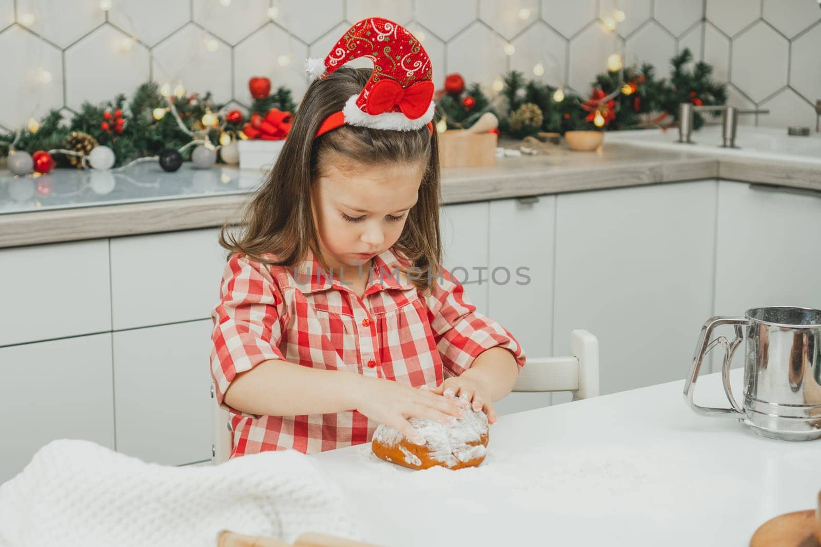 Little dark-haired girl 3 years old in red Christmas cap and checkered shirt prepares dough for gingerbread cookies in white Christmas decorated kitchen with garland lights. Merry Christmas, New Year