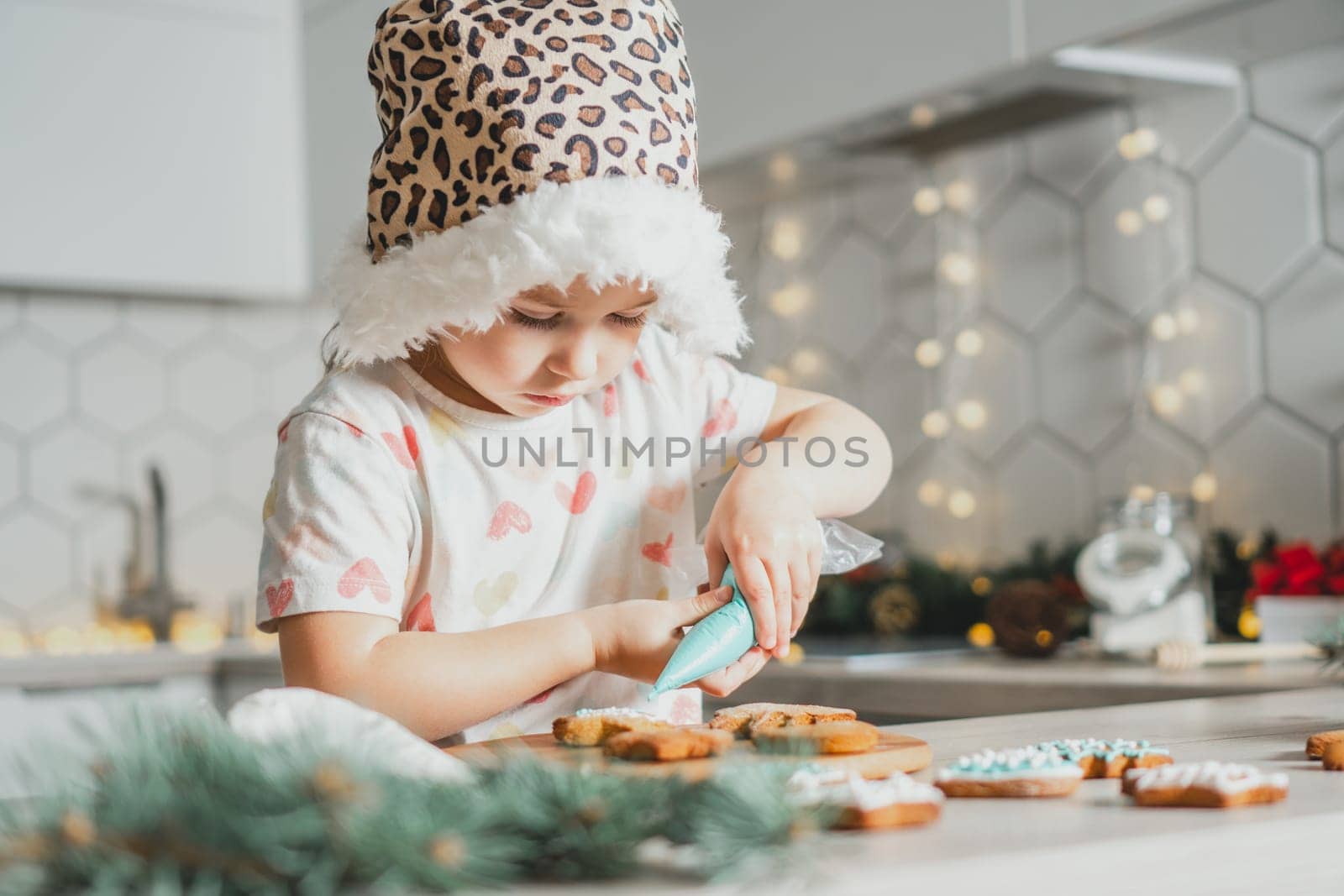 Little dark-haired girl 3 years old in Santa spotted cap decorate gingerbread cookies with icing in white Christmas decorated kitchen. Merry Christmas, Happy New Year