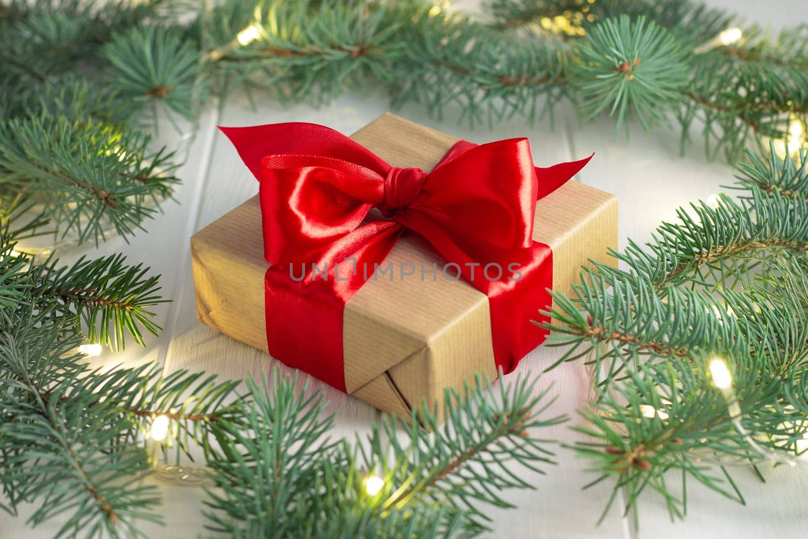 Gift wrapped in craft paper and red ribbon with green branches of a Christmas tree with LED light bulbs garland on a white wooden background. Merry Christmas and Happy New Year concept