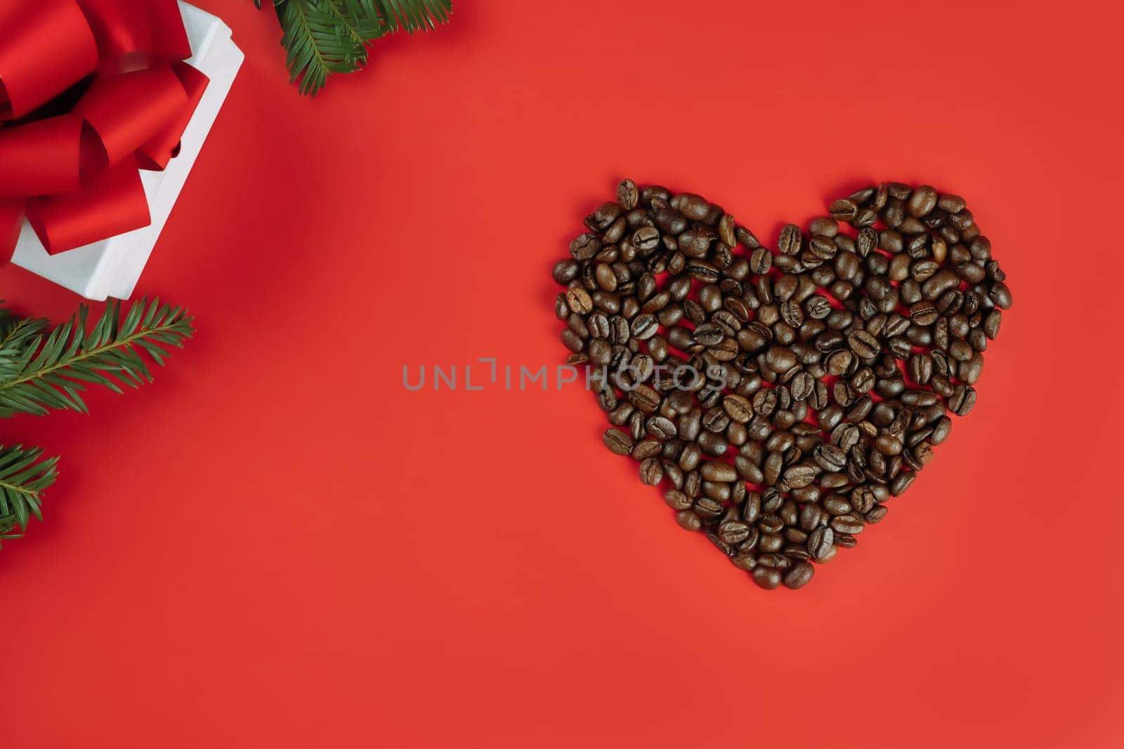 Brown coffee beans laid out in shape of heart with Christmas tree branches and gift box on red background. Flat lay