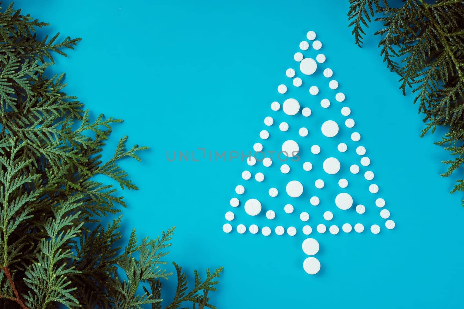White round pills, laid out in shape of Christmas tree with spruce or thuja branches on light blue background. Flat lay. Copy space