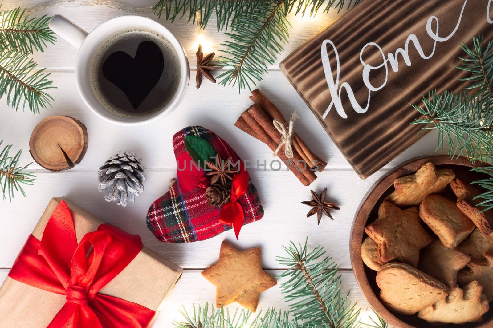 Home lettering burnt wood sign, gingerbread cookie, Christmas tree branches, cup of coffee, gift, cinnamon sticks on white surface. Flat lay