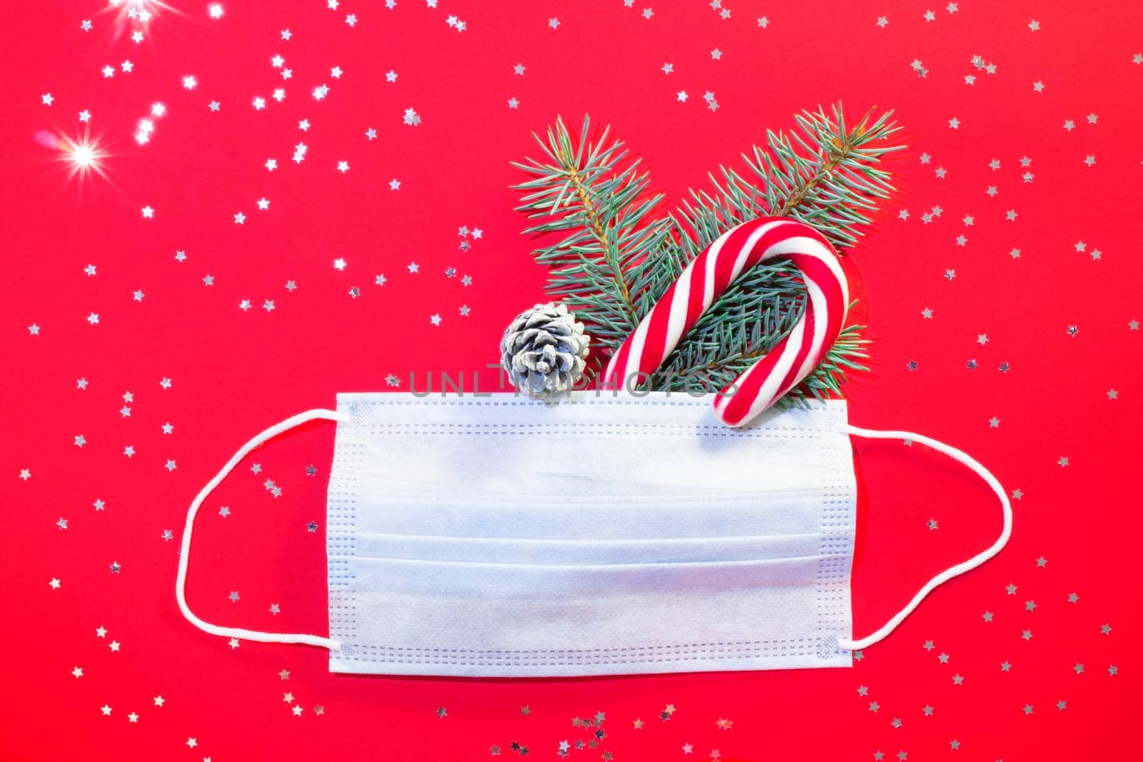 Medical surgical protective mask with Christmas tree branch and lollipop on red background with silver stars. Christmas and New Year during a pandemic concept. Flat lay. Copy space.