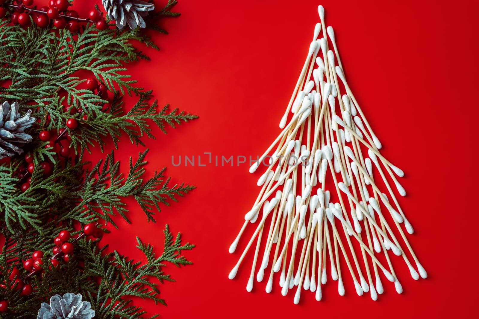 Wooden cotton swabs, laid out in shape of Christmas tree with spruce branches, cones and red berries on red background. Flat lay