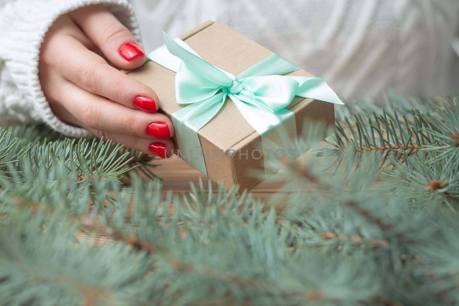 A woman's hand in a white knitted sweater with red nail polish holds a gift with a mint ribbon, near the branches of a Christmas tree.