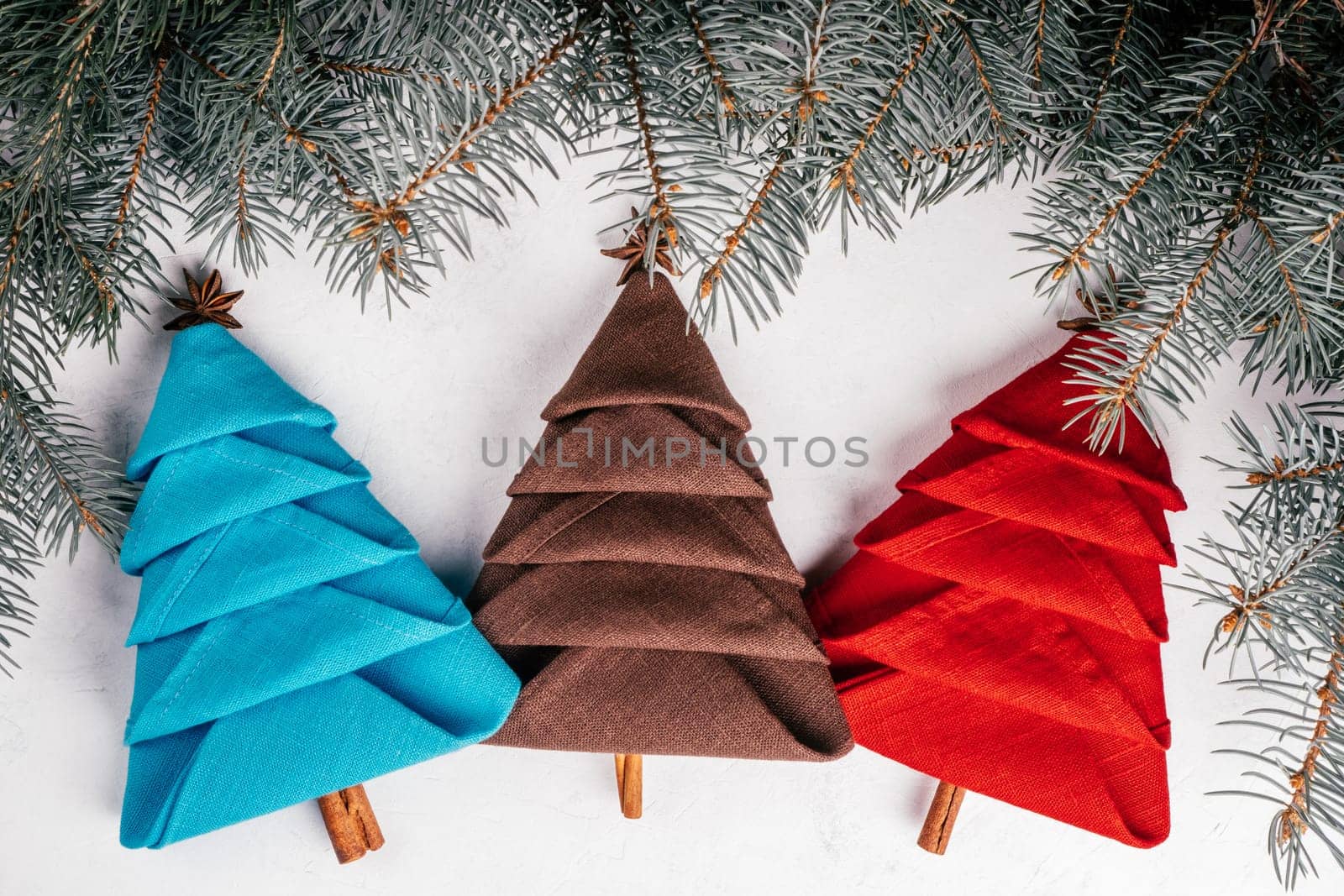Three linen napkins folded in shape of Christmas trees on white concrete surface with spruce branches. New Year table setting. Top view.