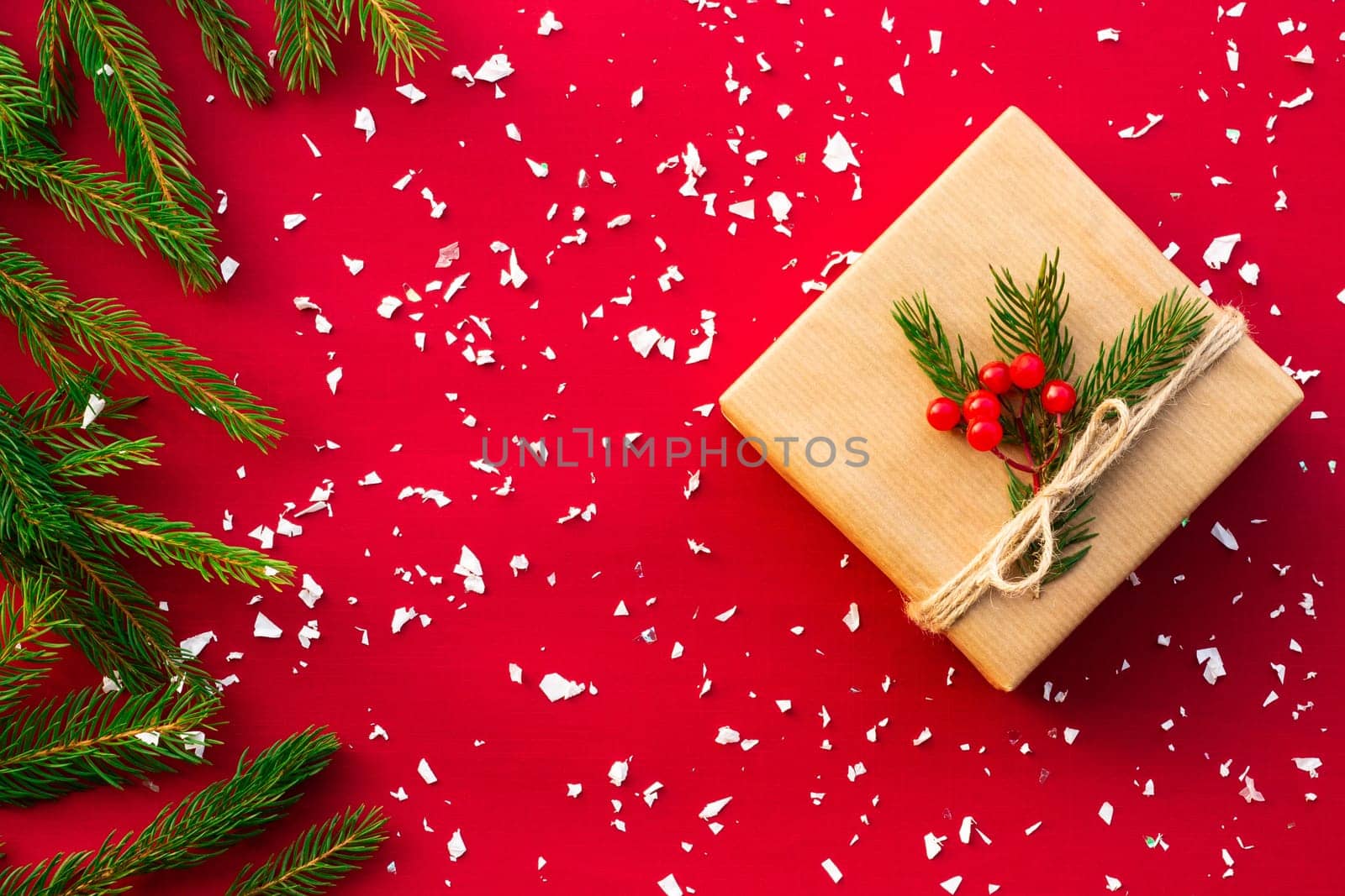 Gift wrapped in craft paper with green branches of a Christmas tree and red berries on a red background. Flat lay. Copy space.