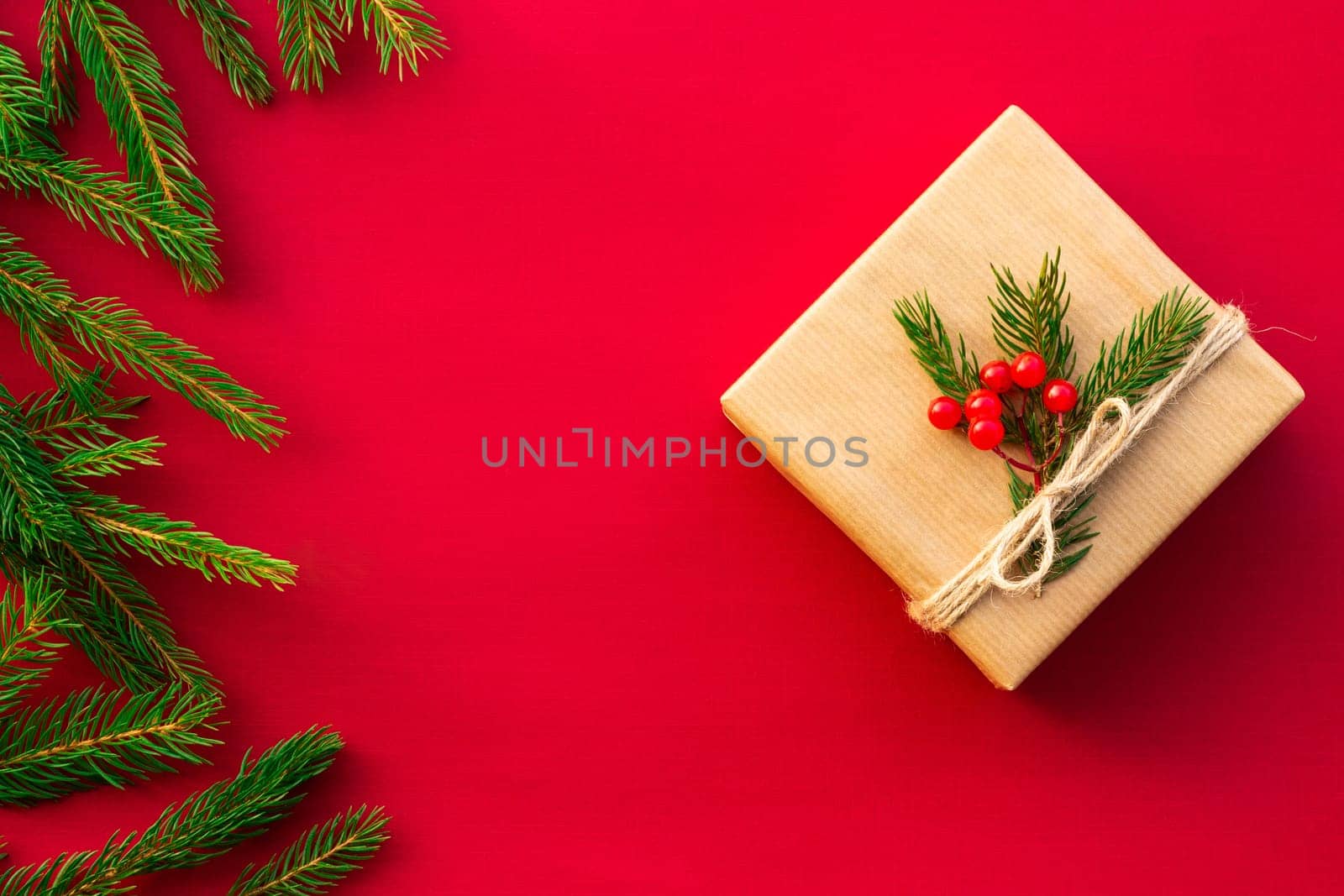 Gift wrapped in craft paper with green branches of a Christmas tree and red berries on a red background. Flat lay. Copy space.