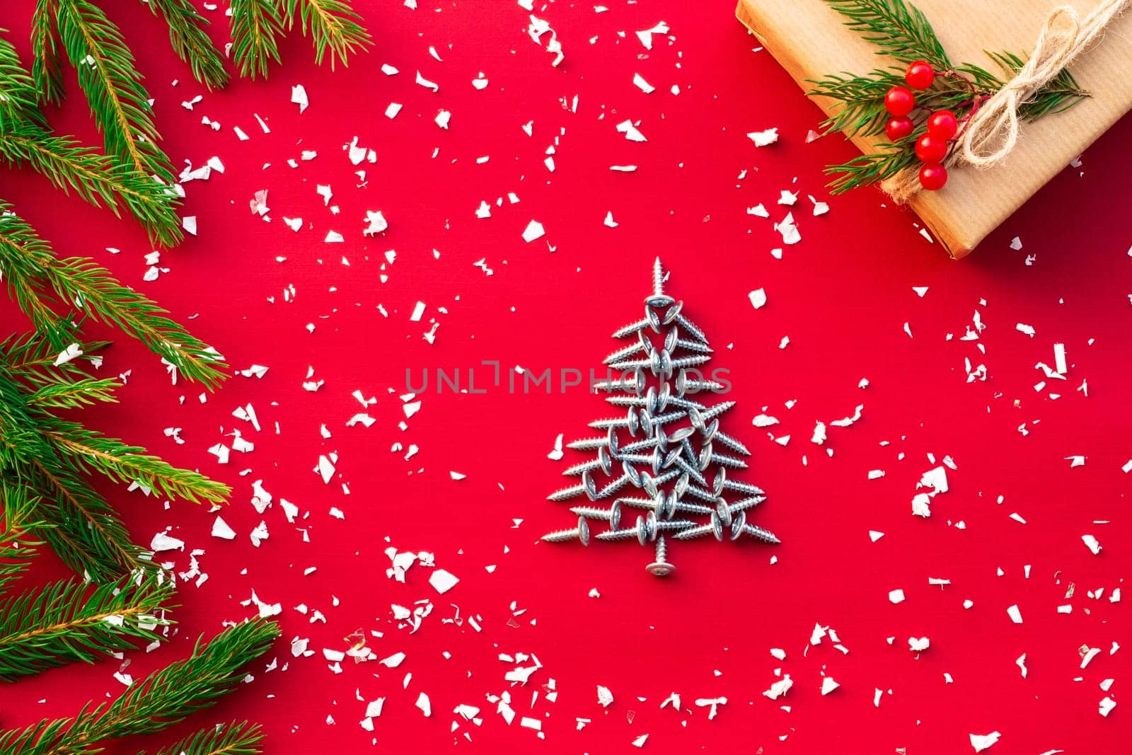 Christmas tree made of self-tapping screws, gift in craft paper with branches of a Christmas tree and red berries on a red background. Flat lay. Copy space.