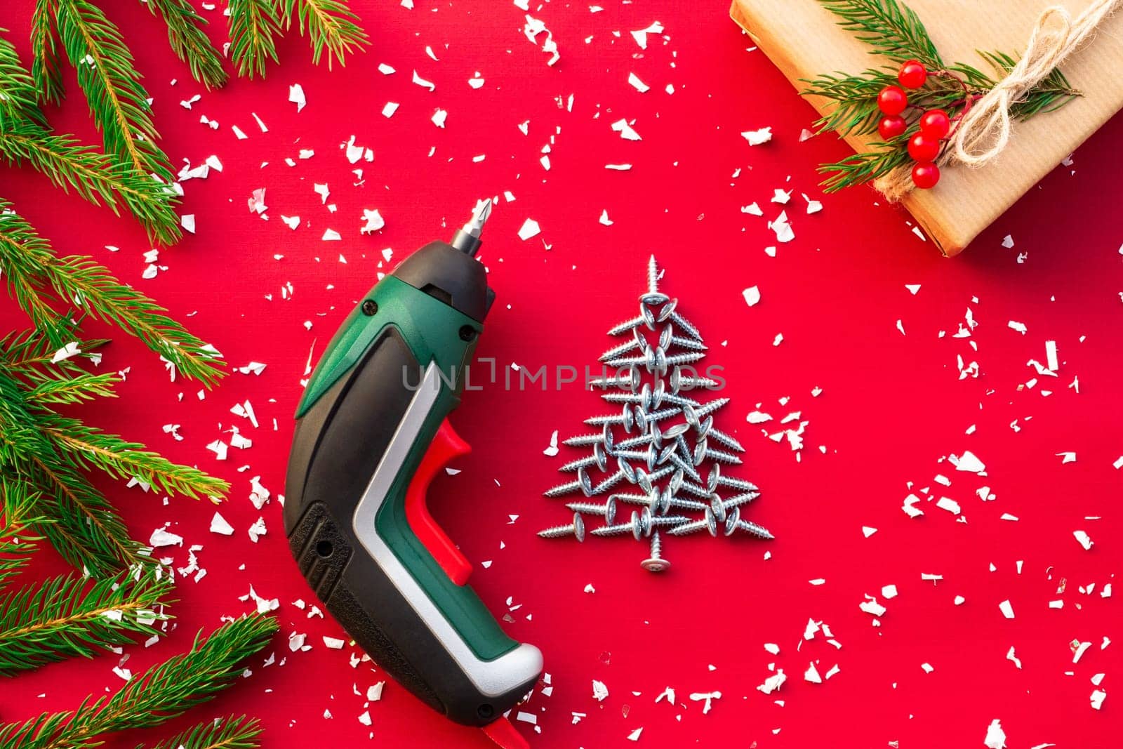 Christmas tree made of self-tapping screws, screwdriver, gift in craft paper with branches of a Christmas tree and red berries on a red background. Flat lay. Copy space.