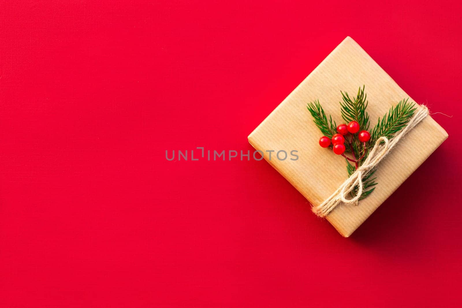 Gift wrapped in craft paper with green branches of a Christmas tree and red berries on a red background. Flat lay. Copy space.