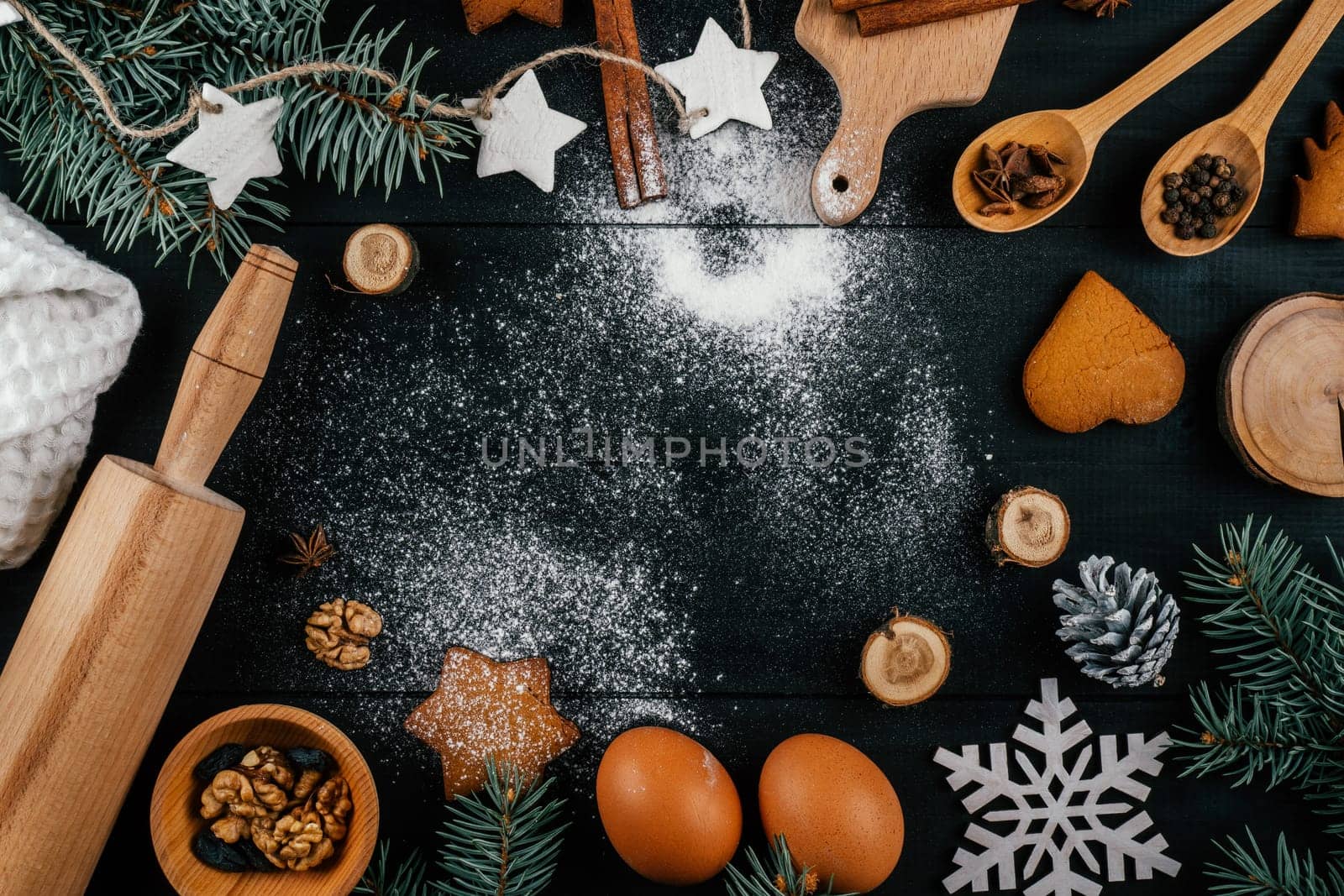 Branches of Christmas tree, gingerbread cookies, baking accessories and ingredients lying on black wooden table with sprinkled flour. Copy space. Merry Christmas and Happy New Year. Top view