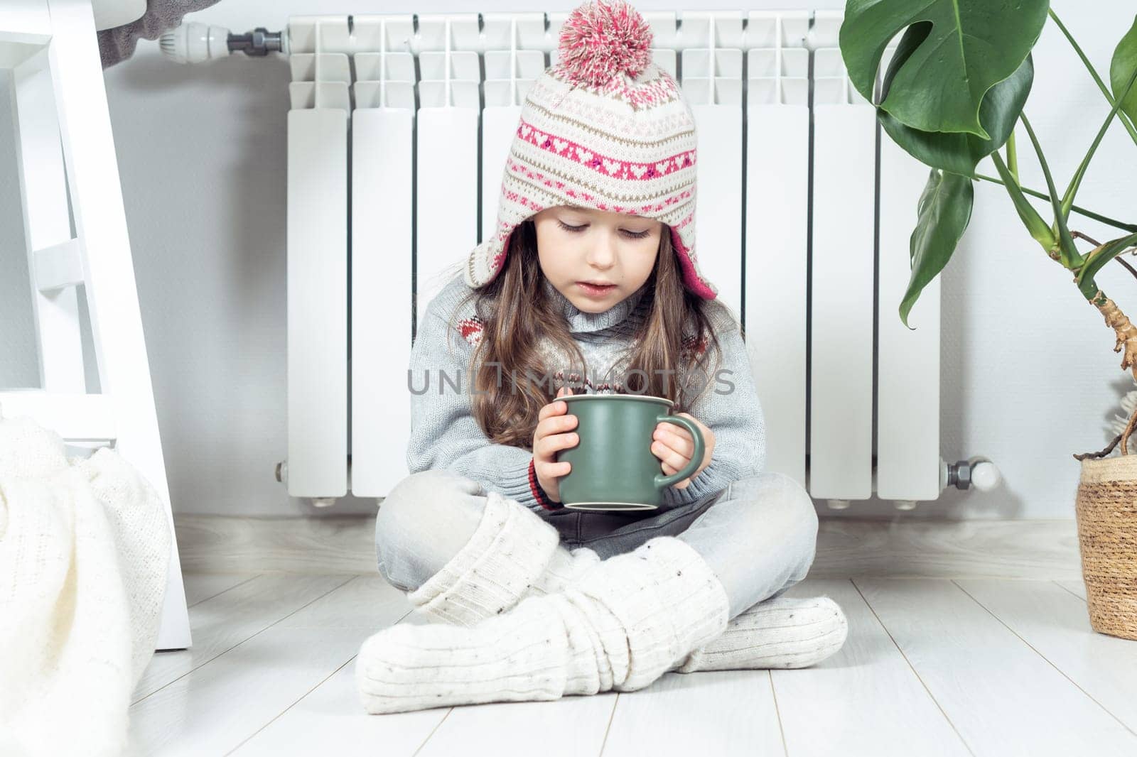 Sad little girl in woolen hat, socks and pullover drink hot tea and sit with crossed legs near heating convector, calorifer. Seasonal sick, virus and flu. Home treatment, healthcare in cold season