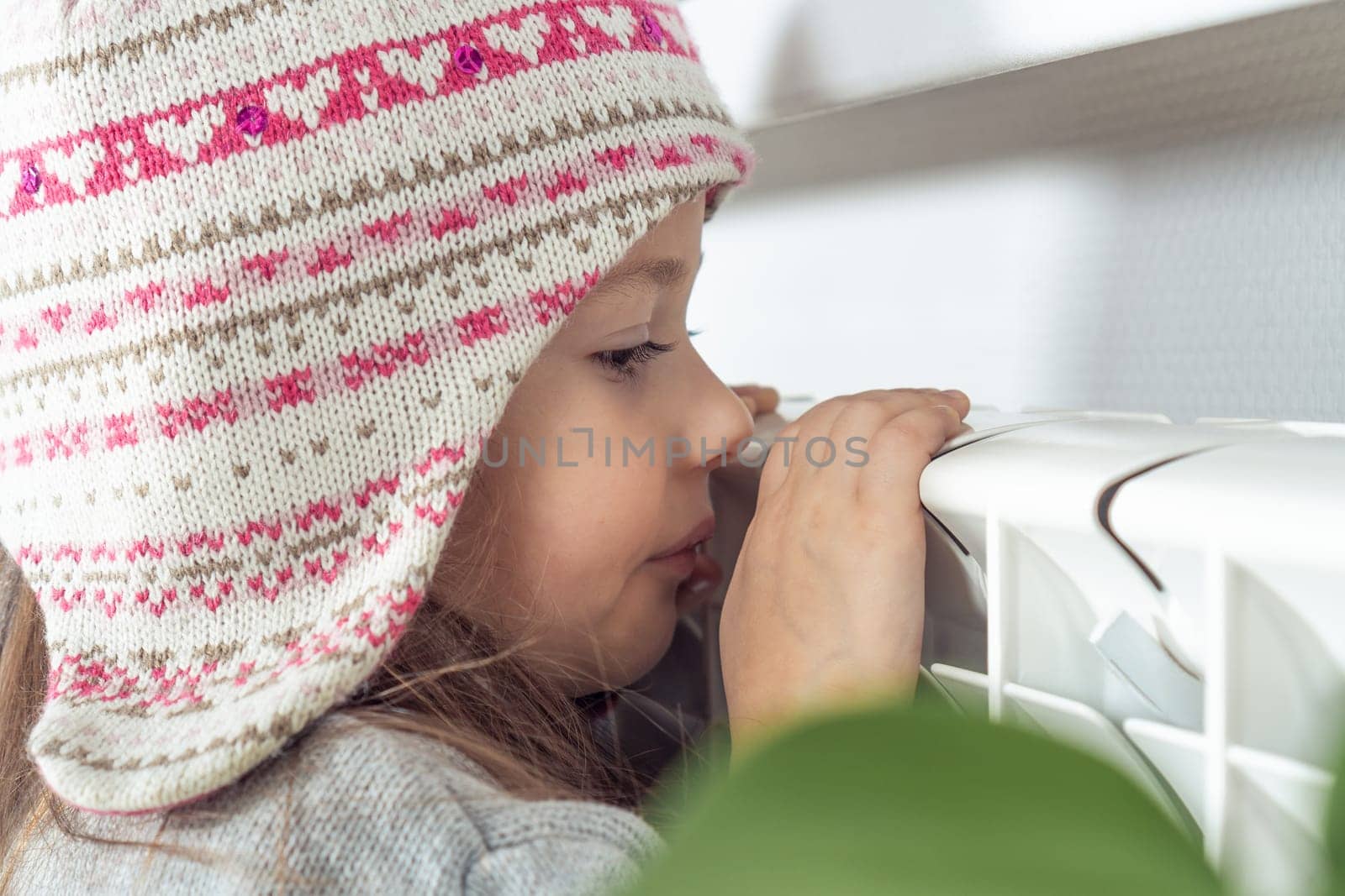 Close up of funny frozen girl face in warm woolen, knitted hat sitting, warming up with nose and hands on heat radiator. Low temperature in apartment. Safety and protection at home. Side view
