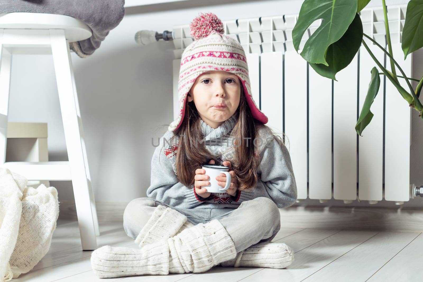 Grimacing little girl in hat, socks and pullover drink sour medicine syrup and sit near heating convector, radiator, next to houseplant. Seasonal sick. Weak throat, immune system in cold winter
