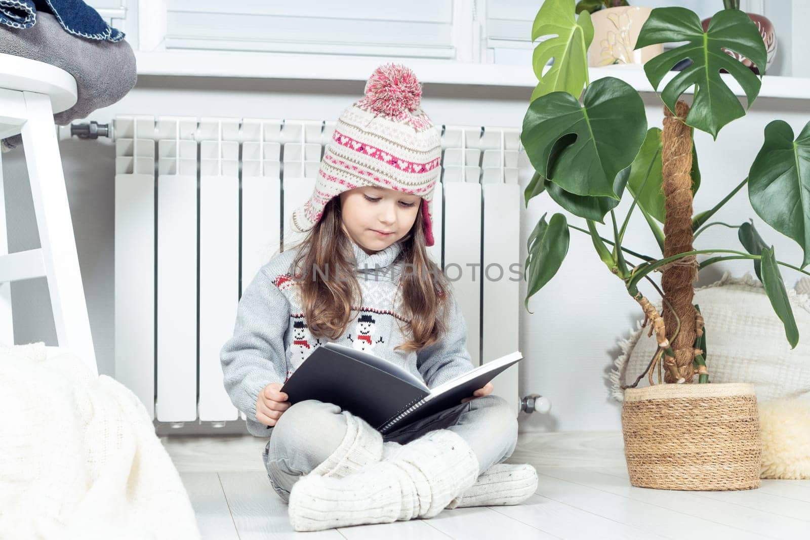 Interested, serious cute little girl in woolen hat and warm clothes with snowman reading book next to heating radiator in white living room. Cozy and comfortable evening with storytelling. Side view