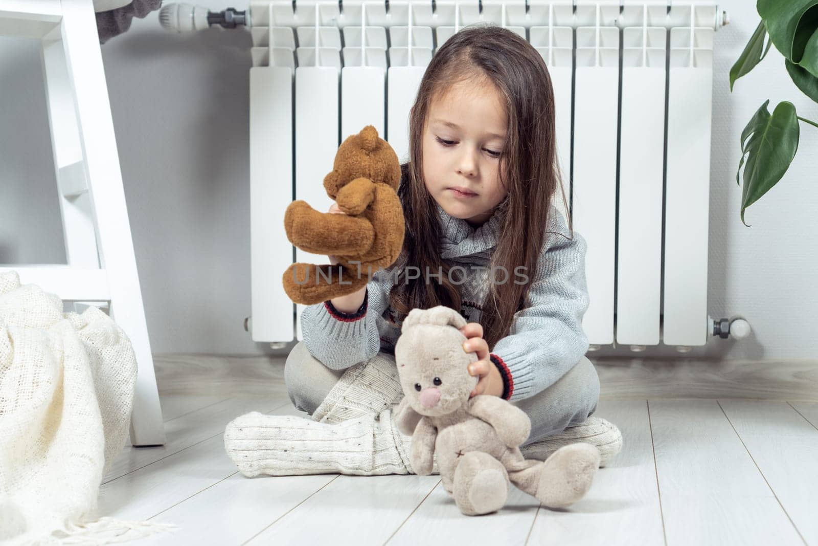 Tranquil adorable little girl sit near heating radiator and playing with stuffed soft bear, rabbit toys, games on the floor, care about imaginary friends. Creative fantasy games and spend time alone
