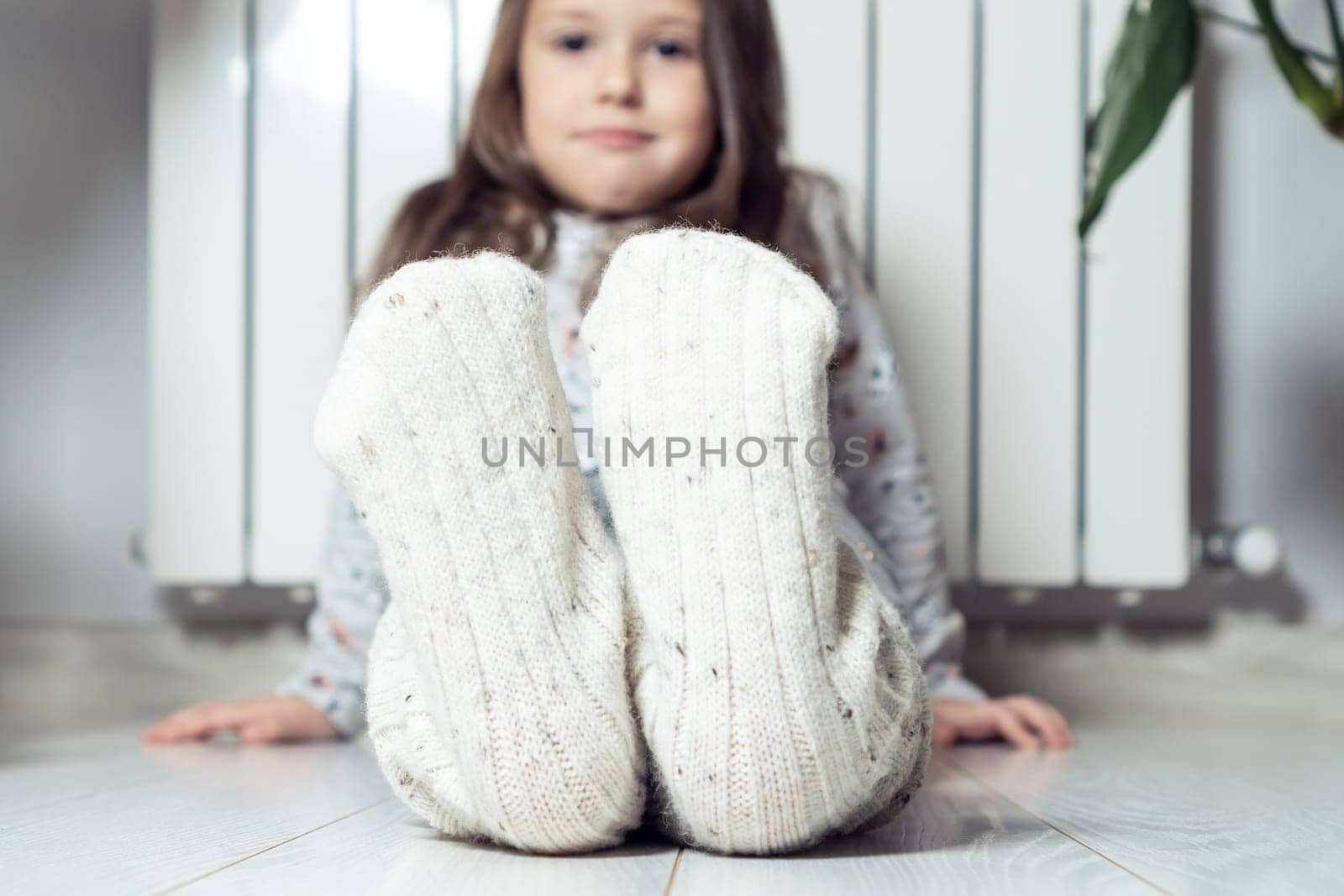 Close up, focus on smiling, calm little girl feet in white woolen socks, showing to camera, sitting near heating radiator at home. Warming up after winter walking, relax. Cozy Christmas holidays