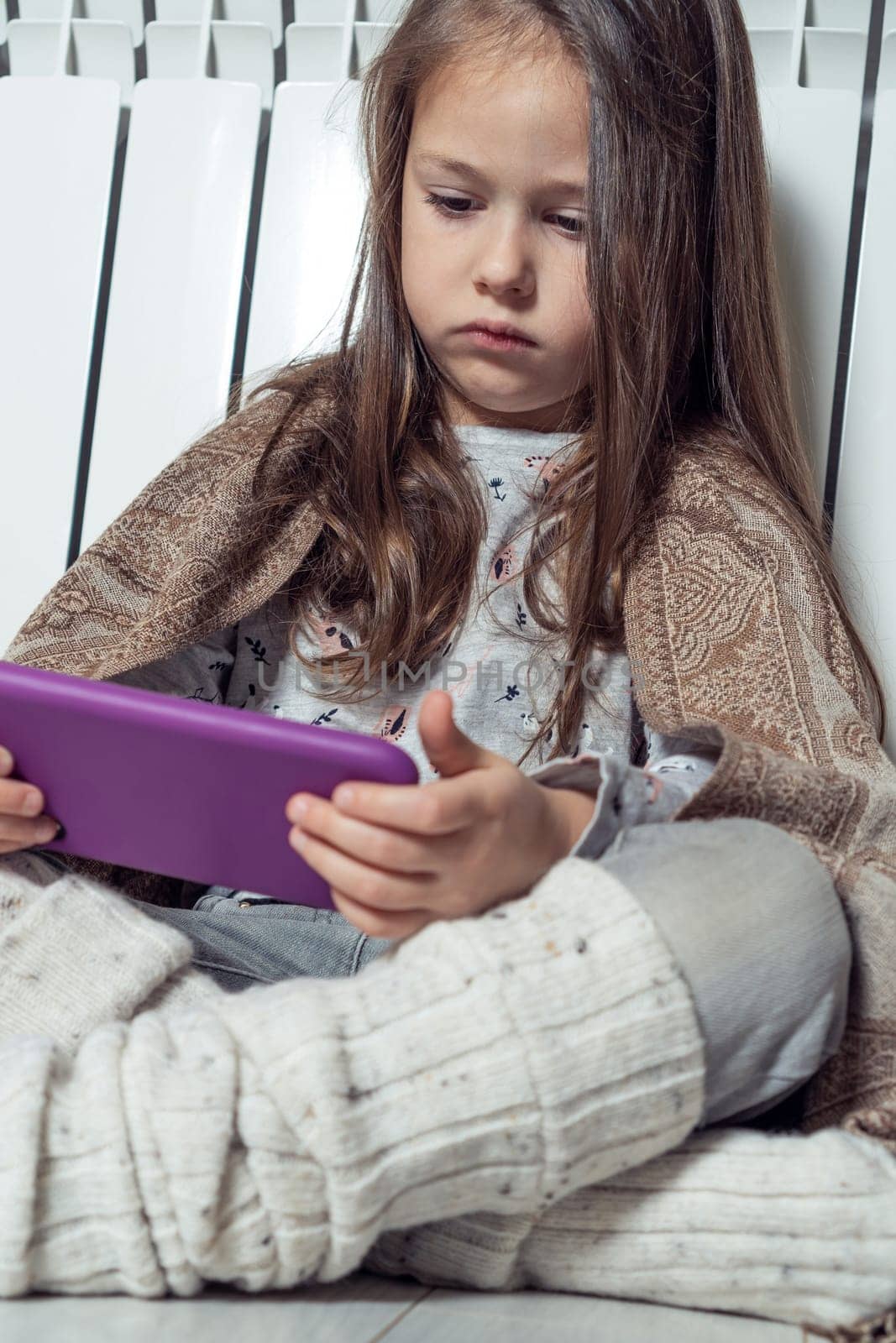Vertical little long haired girl using electronic tablet, playing games and surfing net, wrapped in blanket, sitting near warm heating radiator. Wireless technology and digital native generation