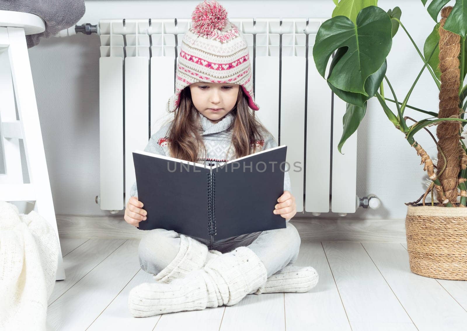Serious, cute, concentrated little girl in knitted hat and warm clothes reading, exploring book next to heating radiator, near plant in white room. Explore and research of notebook or sketchbook