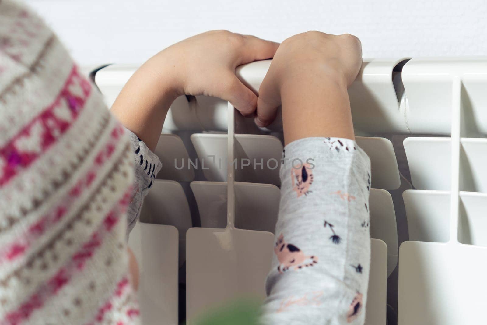 Rearview close up of frozen little child in warm hat and pajamas sitting on knees and warm hands on heat radiator. Safety and protection at home. Climate change and low temperature in apartment