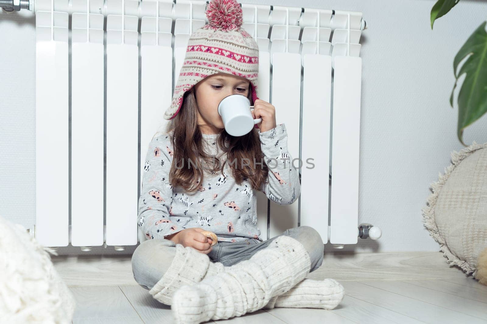 Nice curly little girl in knitted hat and woolen socks eat sweet dessert, drink hot cacao, sitting near white heating radiator in light room. Holidays in winter, hygge and comfort in cold season
