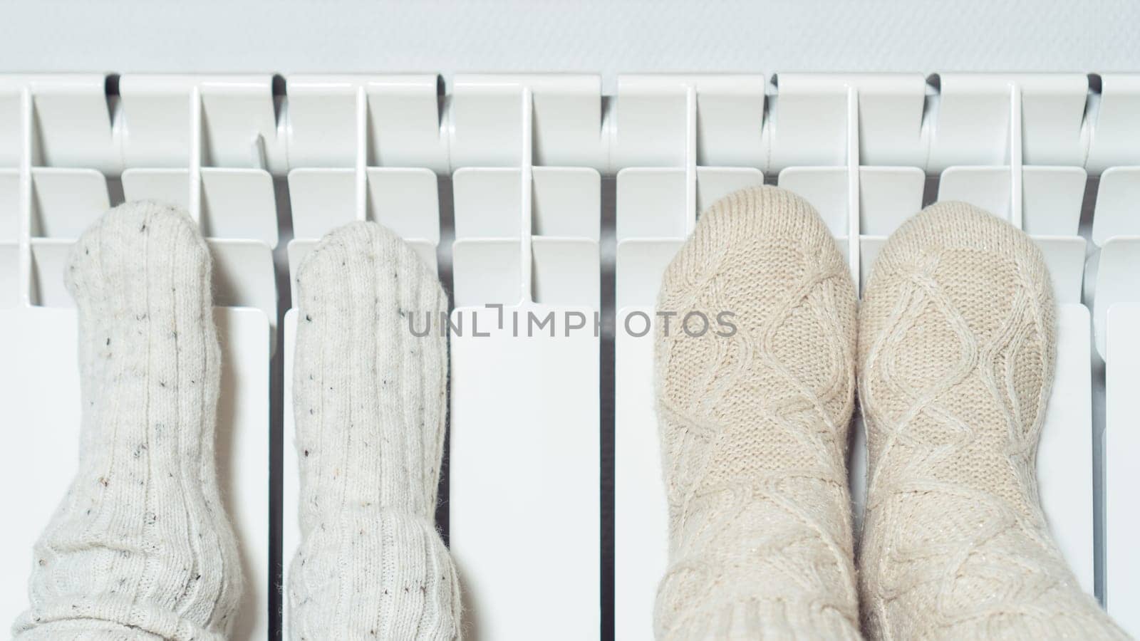Close up photo of cute, cozy child and adult raising legs in wool, knitted socks warm themselves near heat radiator. Central heating, gas consumption in cold winter season. Christmas evening at home