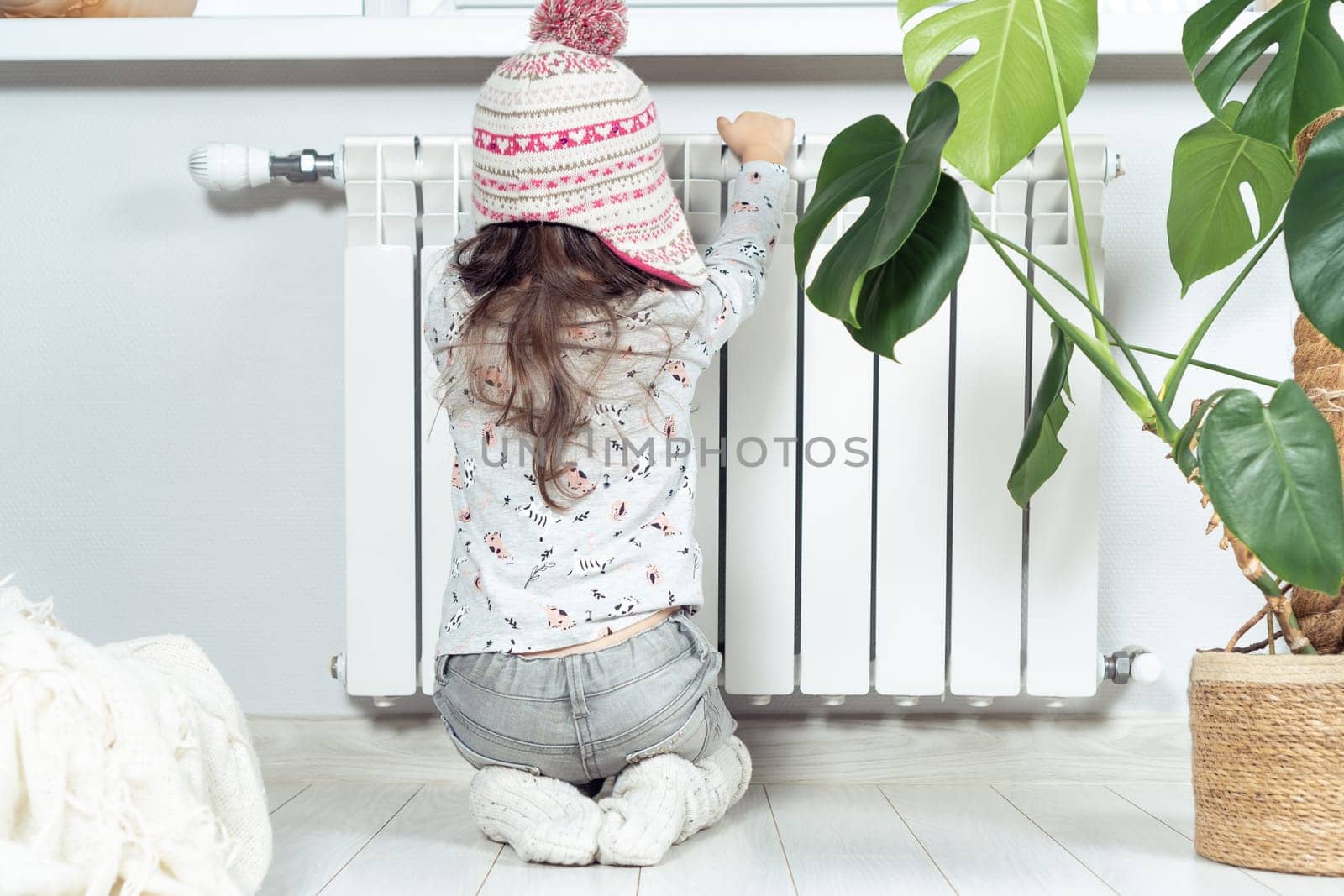 Rearview of frozen little curly girl in warm knitted hat sitting on knees and warm hands on heat radiator. Safety and protection at home. Gas and heating saving. Climate change and low temperature