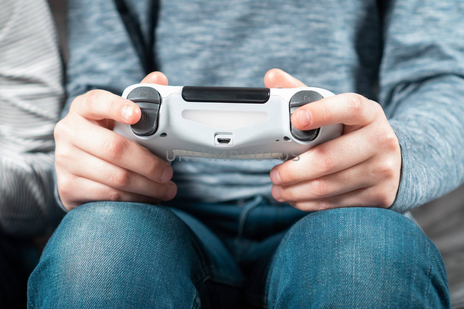 Unrecognizable teenage boy child sitting at home, holding white gaming controller joystick gamepad, playing videogames on game console. Hobby, free time, gaming, interactive entertainment, leisure.