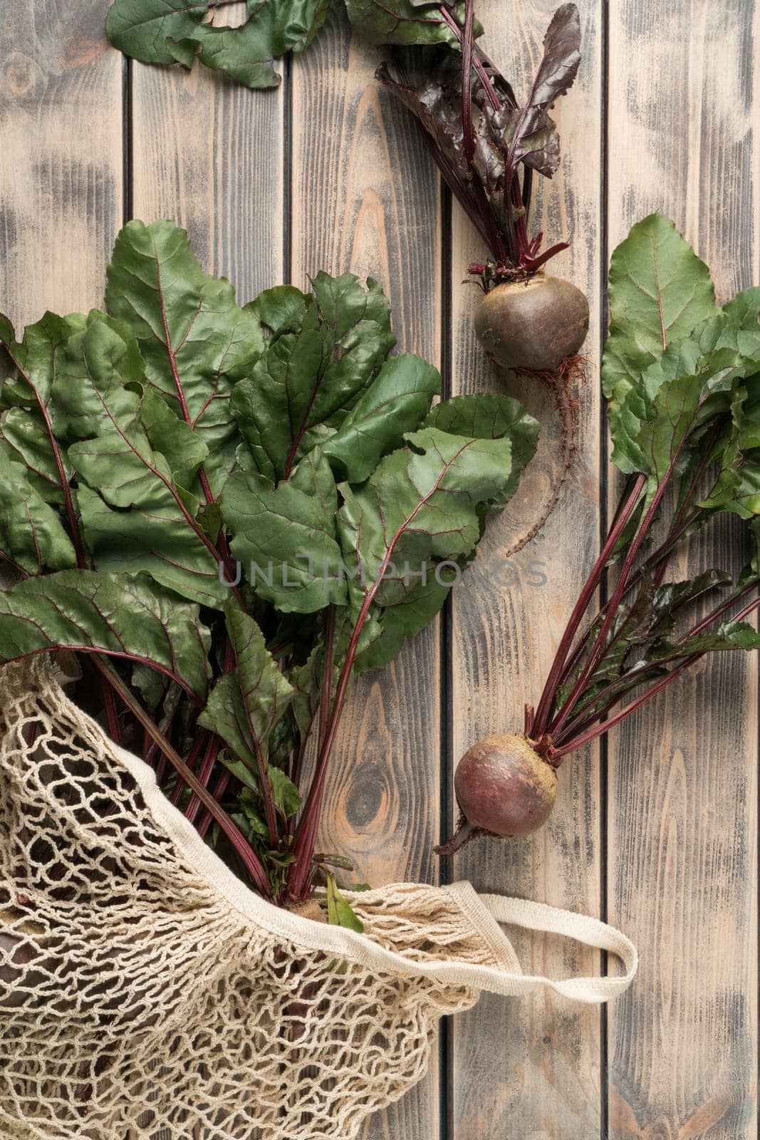 Vertical set of raw fresh organic rural beetroots with stems and leaves in string bag on wooden background. Ecological shopping with natural bags, sustainable development. Top view, copy space