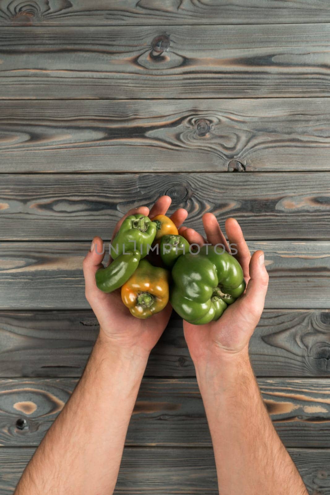 Vertical close up shot of cropped man hands holding fresh, juicy green bell pepper vegetable on wooden table background. Rustic freshness, agriculture season. Top view, above, copy space, mockup