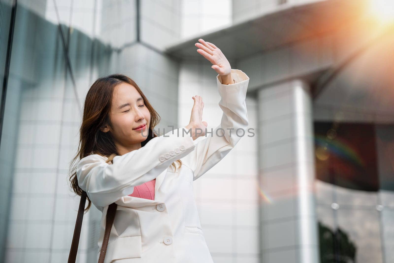 Happy woman wearing white suit jacket with brown bag at sunlight outdoors. Portrait of confidence young businesswoman standing outside office building in city raise your hand to shade sun