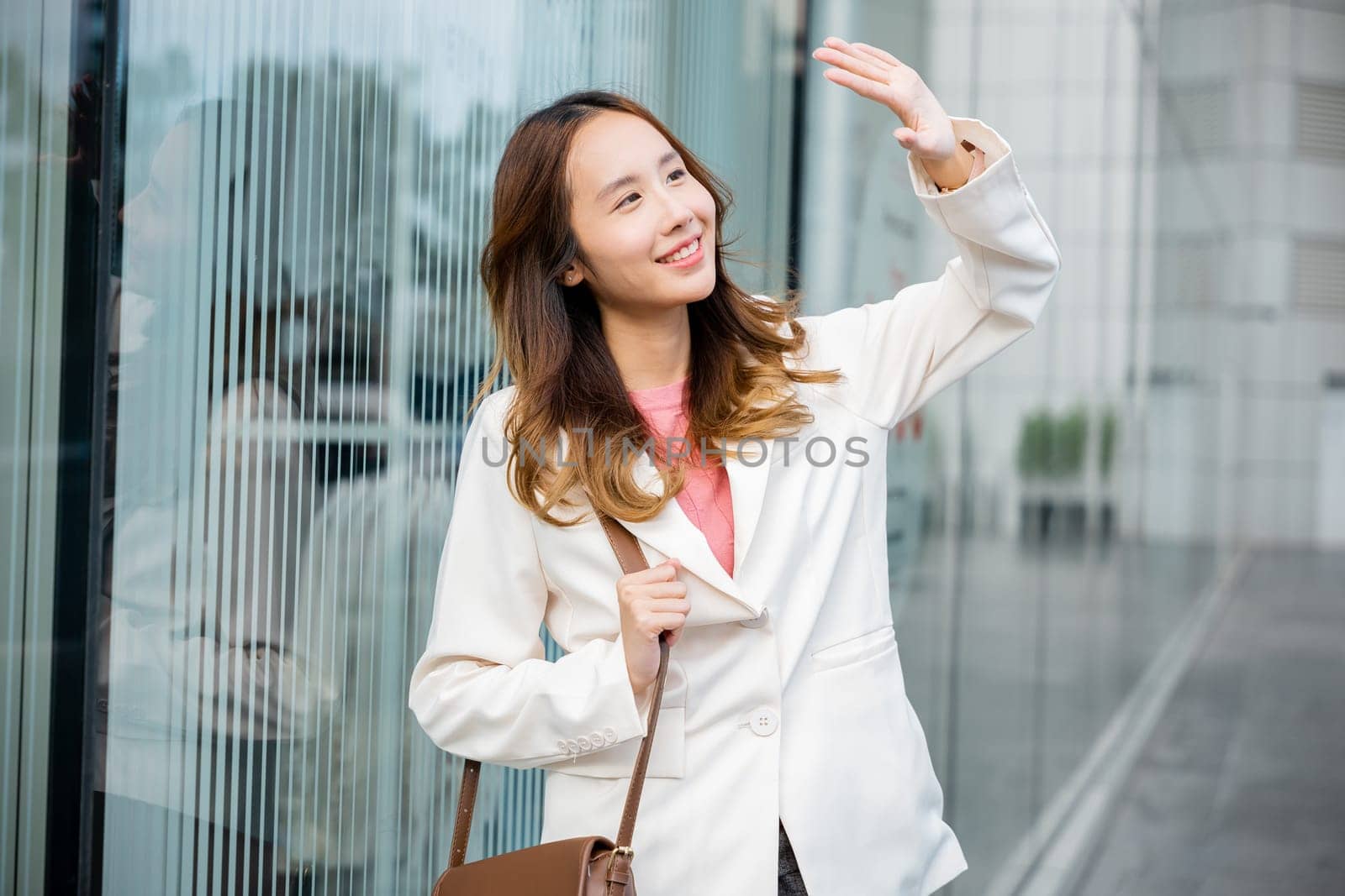 Portrait of confidence young businesswoman standing outside office building in city. Happy woman wearing white suit jacket with brown bag at sunlight outdoors.