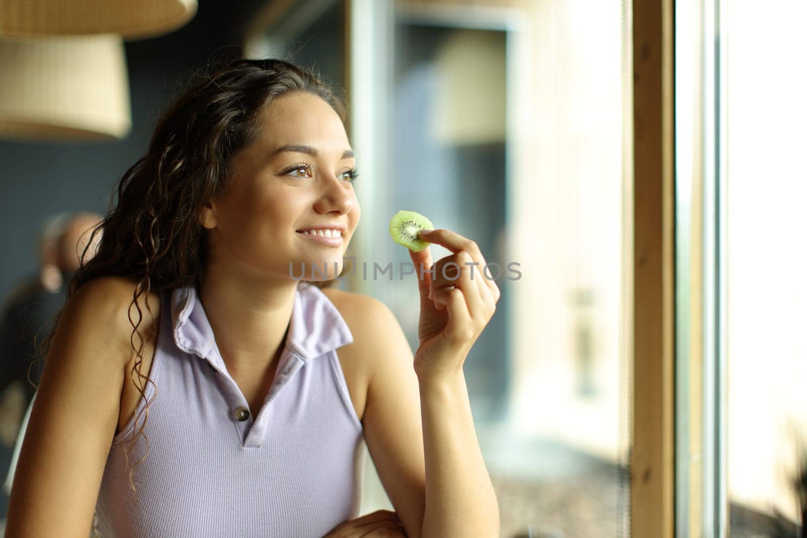 Happy woman in a restaurant eating kiwi looking away through a window