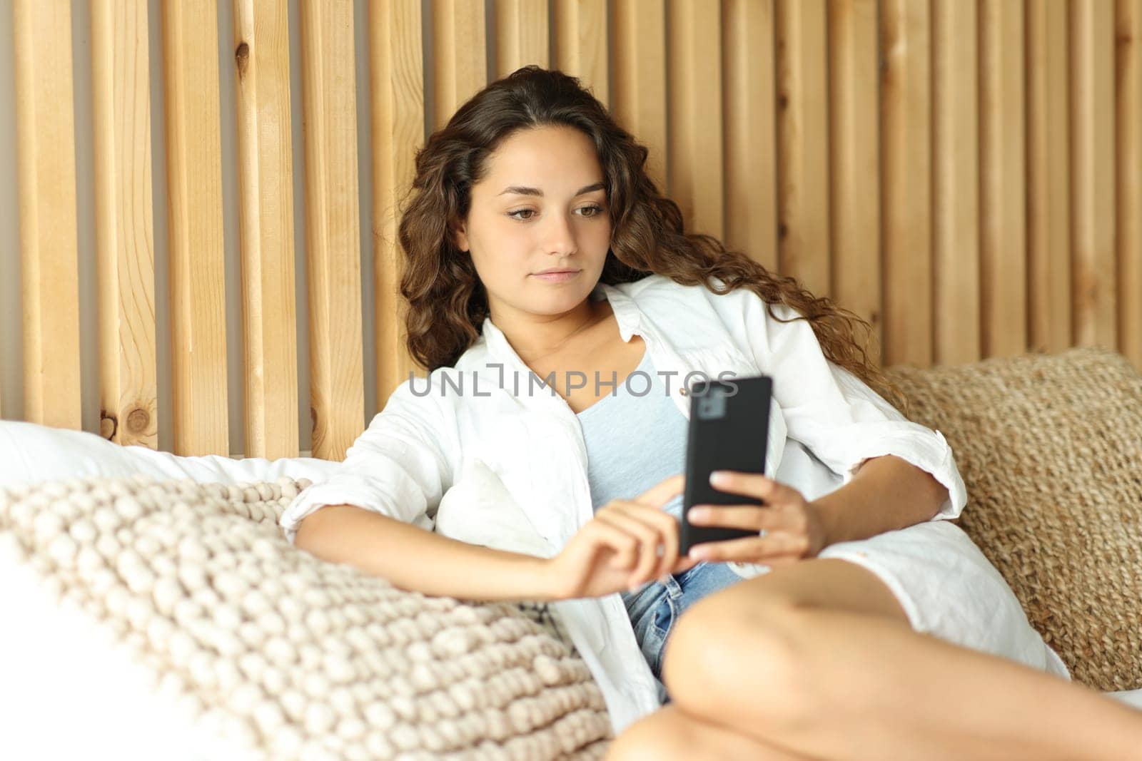 Serious woman using mobile phone sitting on a bed in a room at home