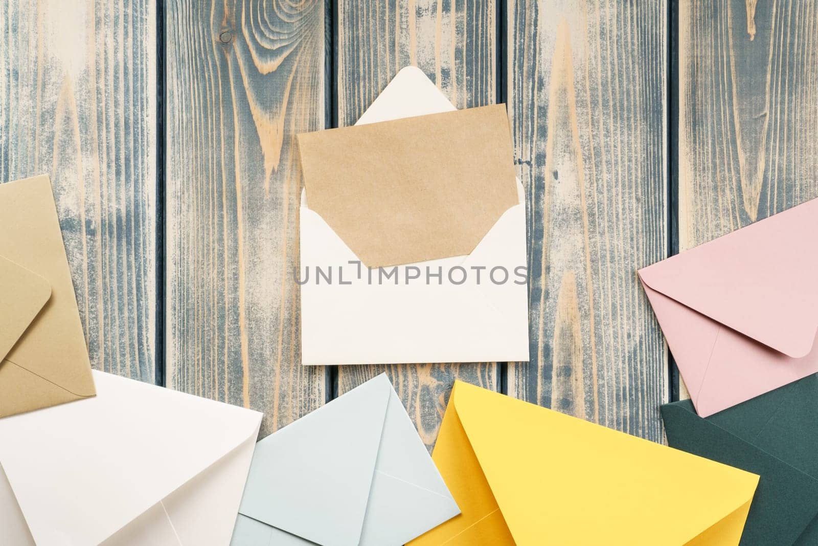 White open envelope with open kraft paper card on gray wooden background. Multicolored envelopes lie side by side. Copy space. Top view. Mail, correspondence, sending messages, congratulations