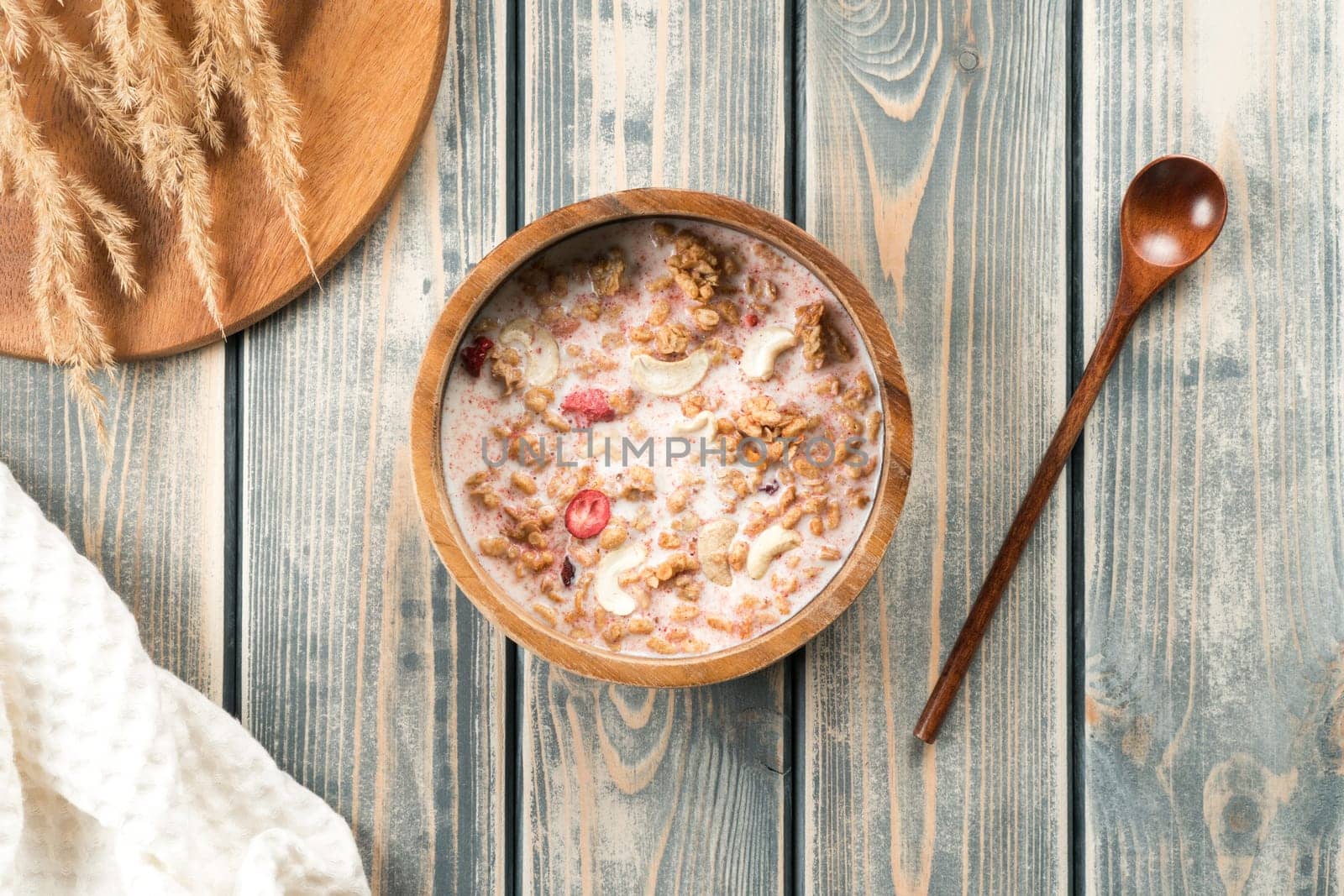 Wooden bowl filled with whole grain granola with berries and nuts poured with milk. Delicious and healthy breakfast. Tray of spikelets, spoon and white towel on rustic wooden surface. Top view