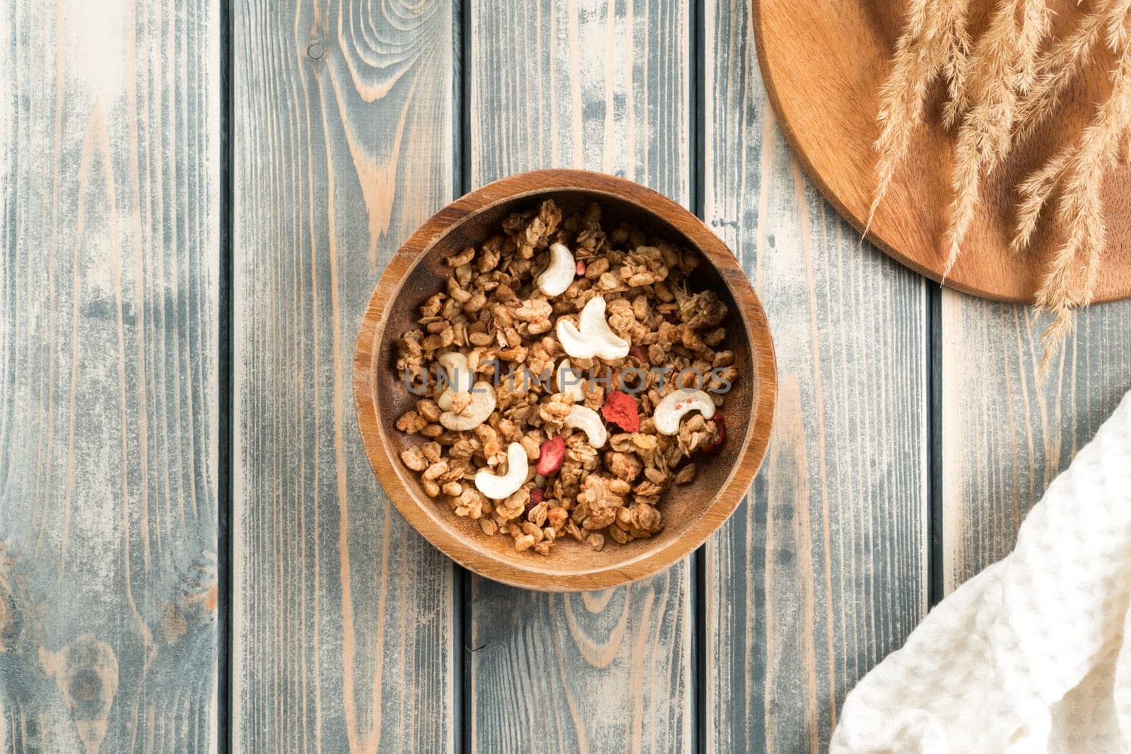 Wooden bowl filled with whole grain granola with berries and nuts. Delicious and healthy breakfast. Tray of spikelets and white towel on rustic wooden surface. Top view