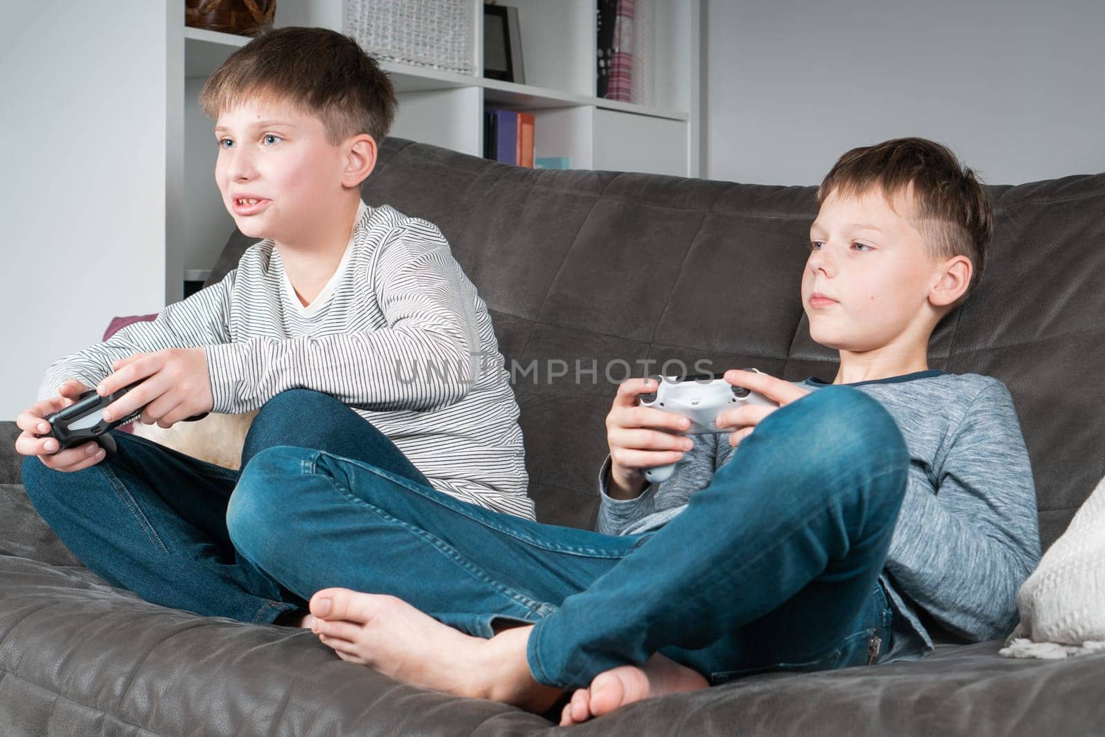 Portrait of two concentrated barefoot teenage boys sitting on grey sofa at home, holding gaming controller joystick, playing videogames. Hobby, free time, gaming, interactive entertainment, leisure.