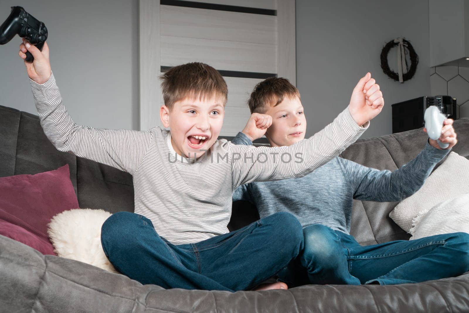 Portrait of two joyful cheerful teenage boys sitting on grey sofa at home, holding gaming controller joystick, playing videogames, celebrating, raising fists, winning competition. Hobby, free time.