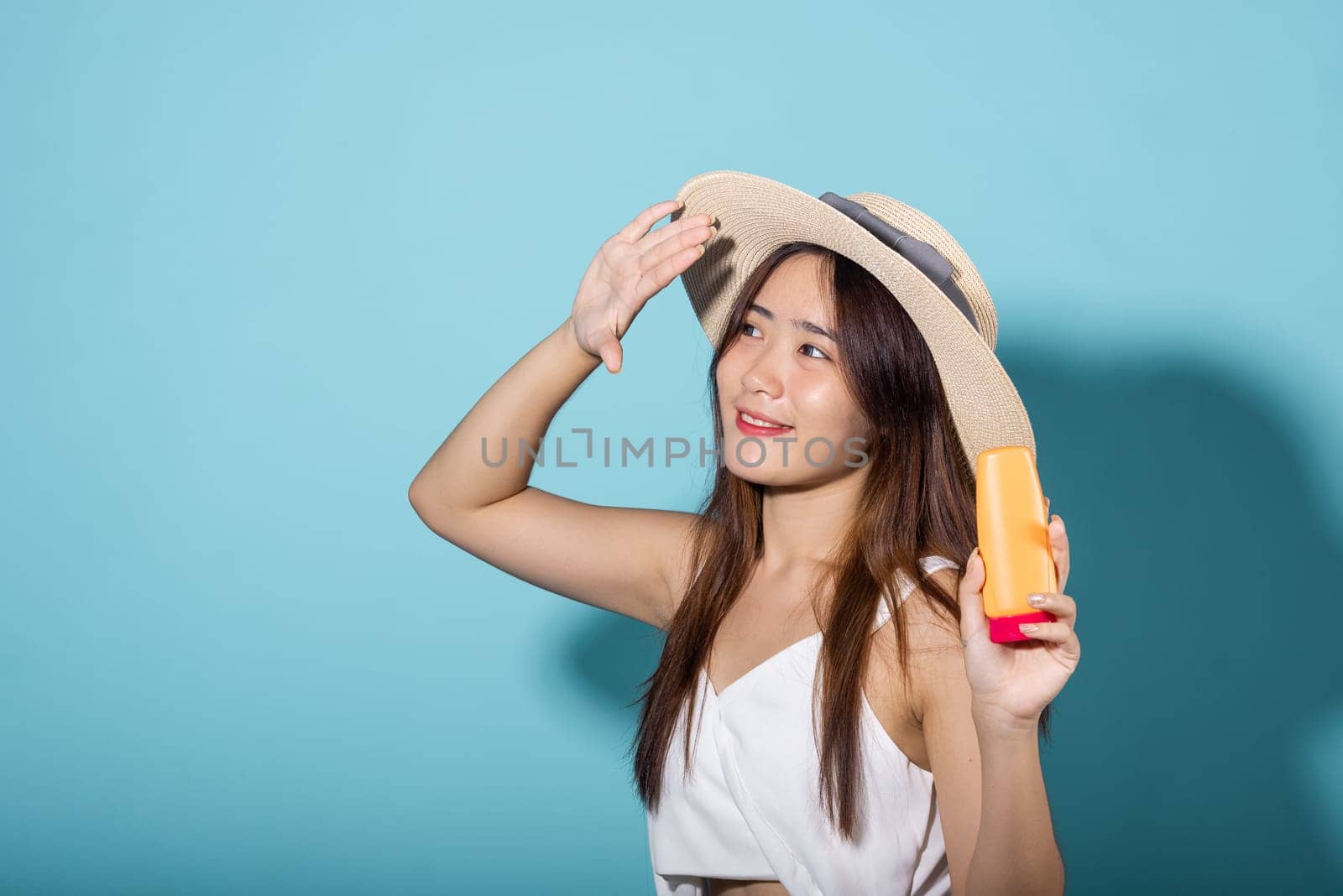 Beautiful Asian young woman holding body lotion on hand in studio shot isolated on blue background, Smiling brunette female hold cream sunblock bottle, tube of sunscreen, protect sun light and UV