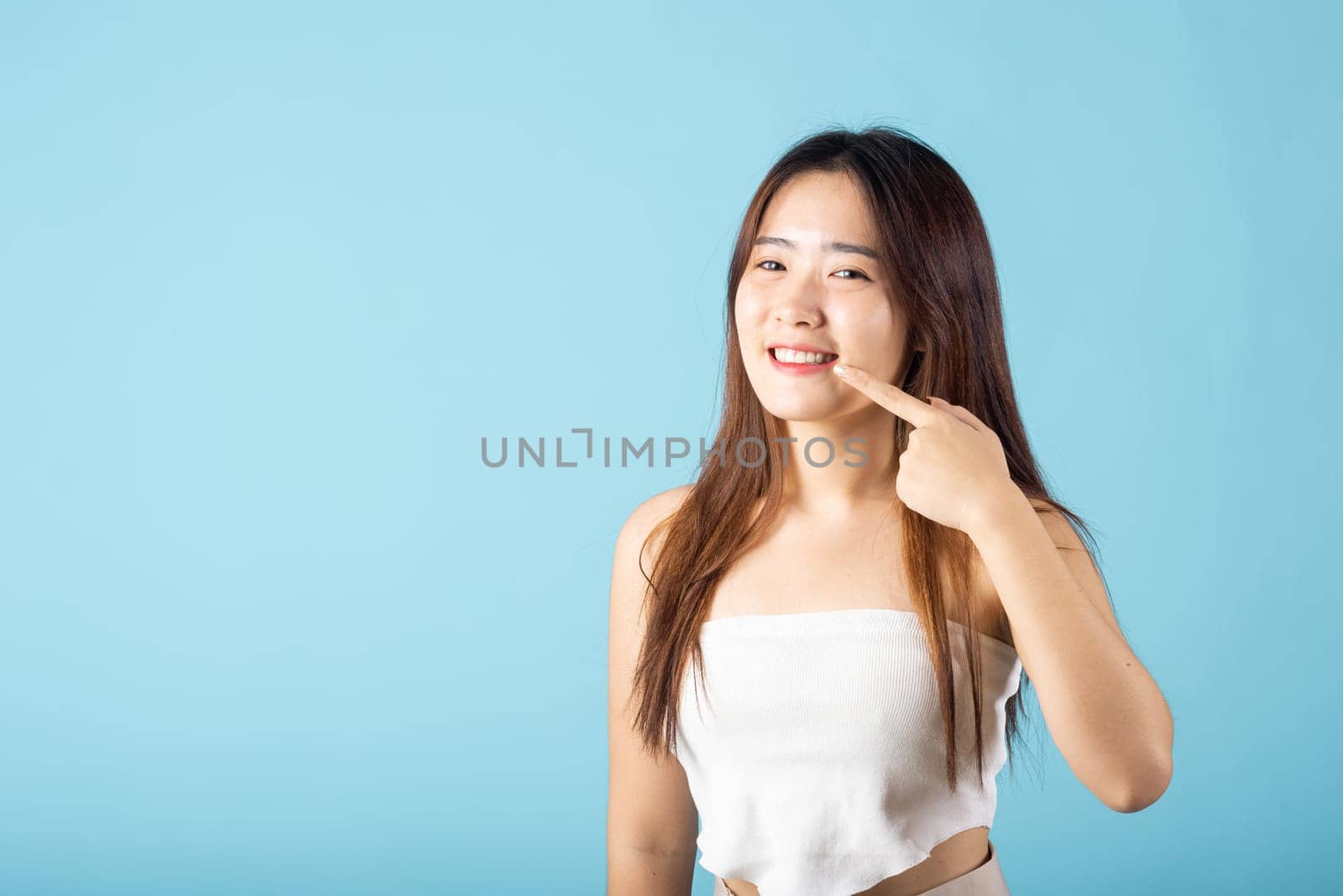Dental health. Asian young woman pointing to her strong white teeth studio shot isolated on blue background, beautiful female point fingers to smiling mouth