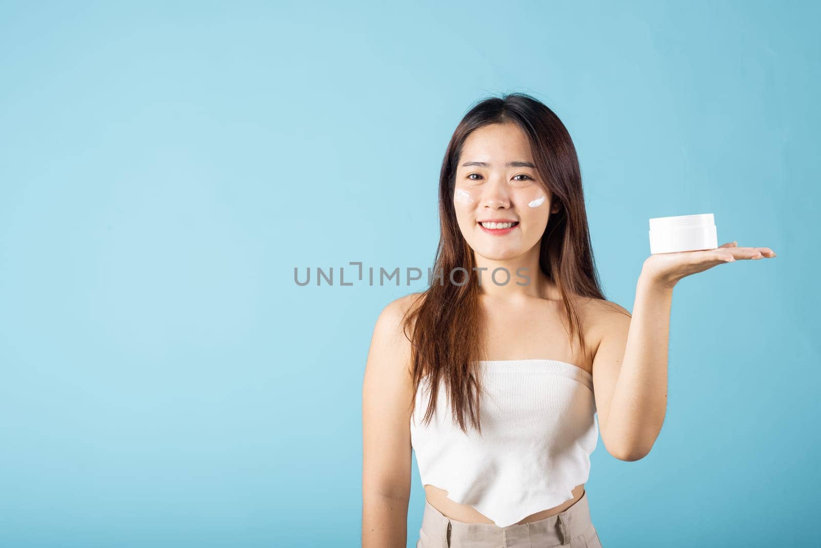 Asian young woman are not very beautiful hold jar of skin cream for face and apply anti-aging moisturizer studio isolated on blue background, Happy female pointing finger her face, pursuit of beauty